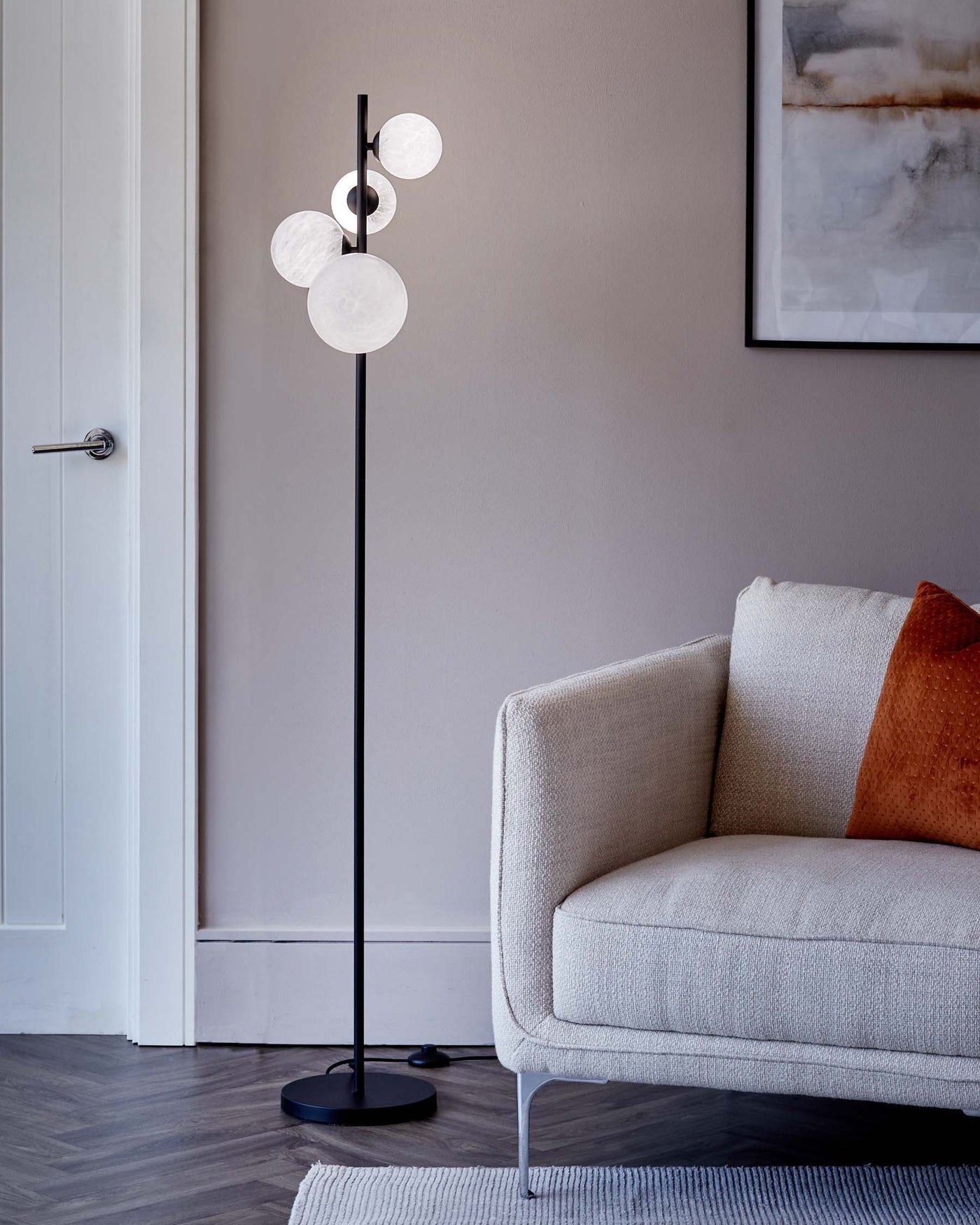 Modern minimalist style furniture in a living room setting, featuring a light grey fabric sofa with a sleek design and slim metal legs. Next to it stands a contemporary black floor lamp with a unique design of multiple frosted glass globes at varying heights on a slender vertical rod. A small textured pillow adds a pop of orange colour to the sofa, and under both pieces rests a light grey woven area rug on dark hardwood flooring. The decor is complemented by a framed abstract wall painting in soft hues.