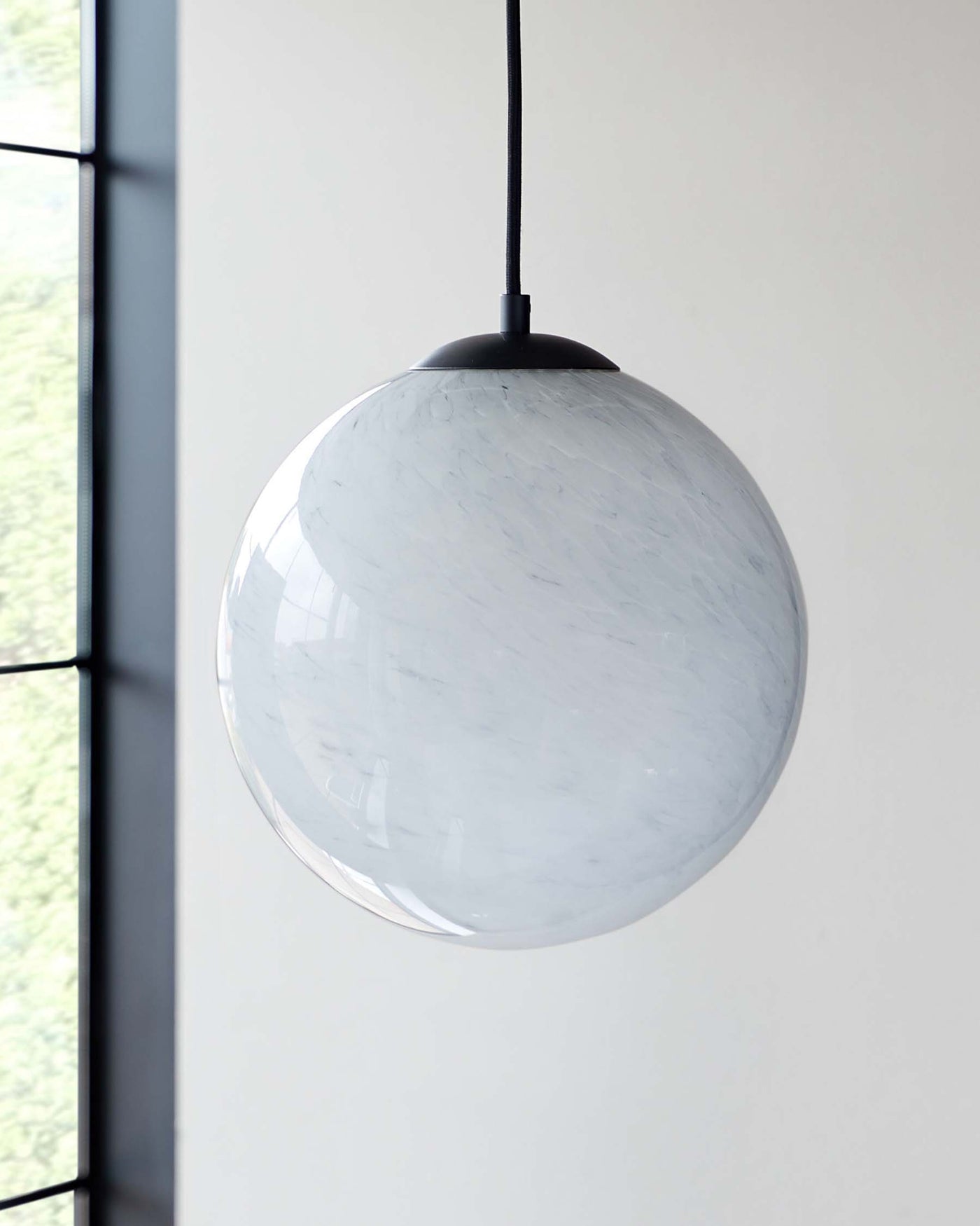 A modern spherical pendant light with a white marble pattern, suspended by a black cord against a neutral background with large windows giving a glimpse of greenery outside.