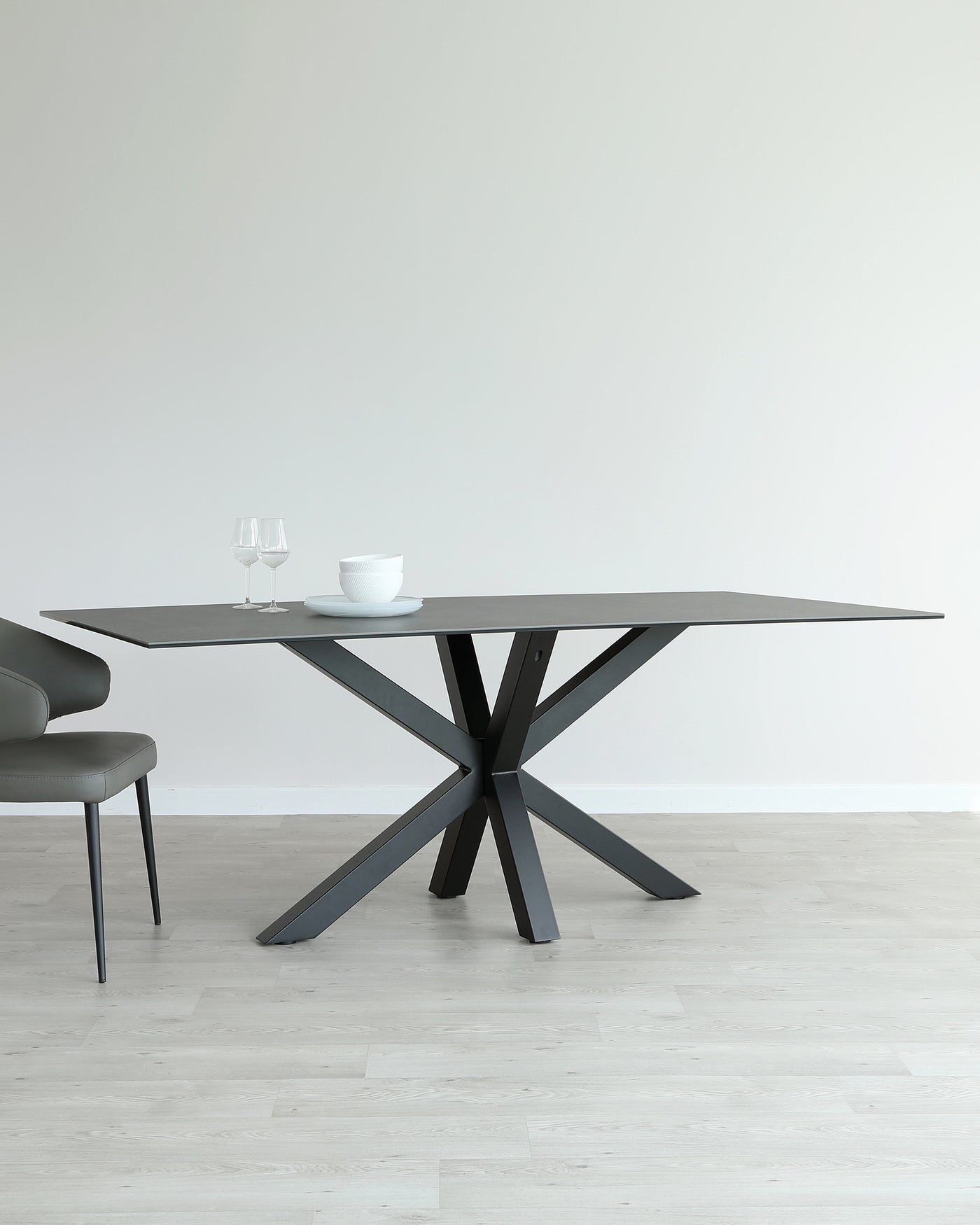 Modern minimalist dining set including a rectangular dark table with an angled cross-leg design and a matching curved bench-style seat with slender legs, set on a light wood floor against a clean white wall, adorned with simple white dinnerware and clear wine glasses.