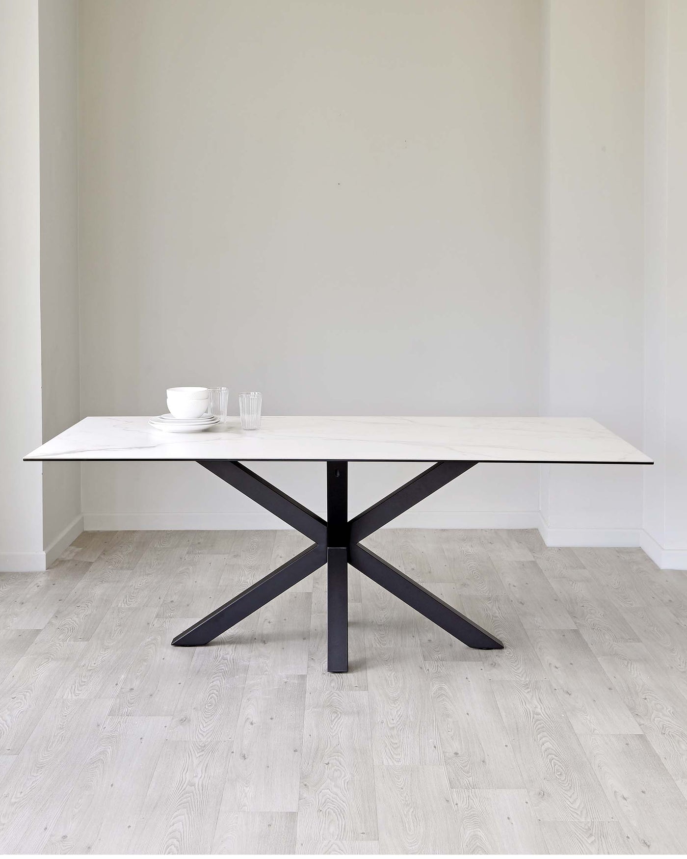 Modern minimalist dining table with a white marble top and a black criss-cross steel base, set in a room with light wooden flooring and pale grey walls.