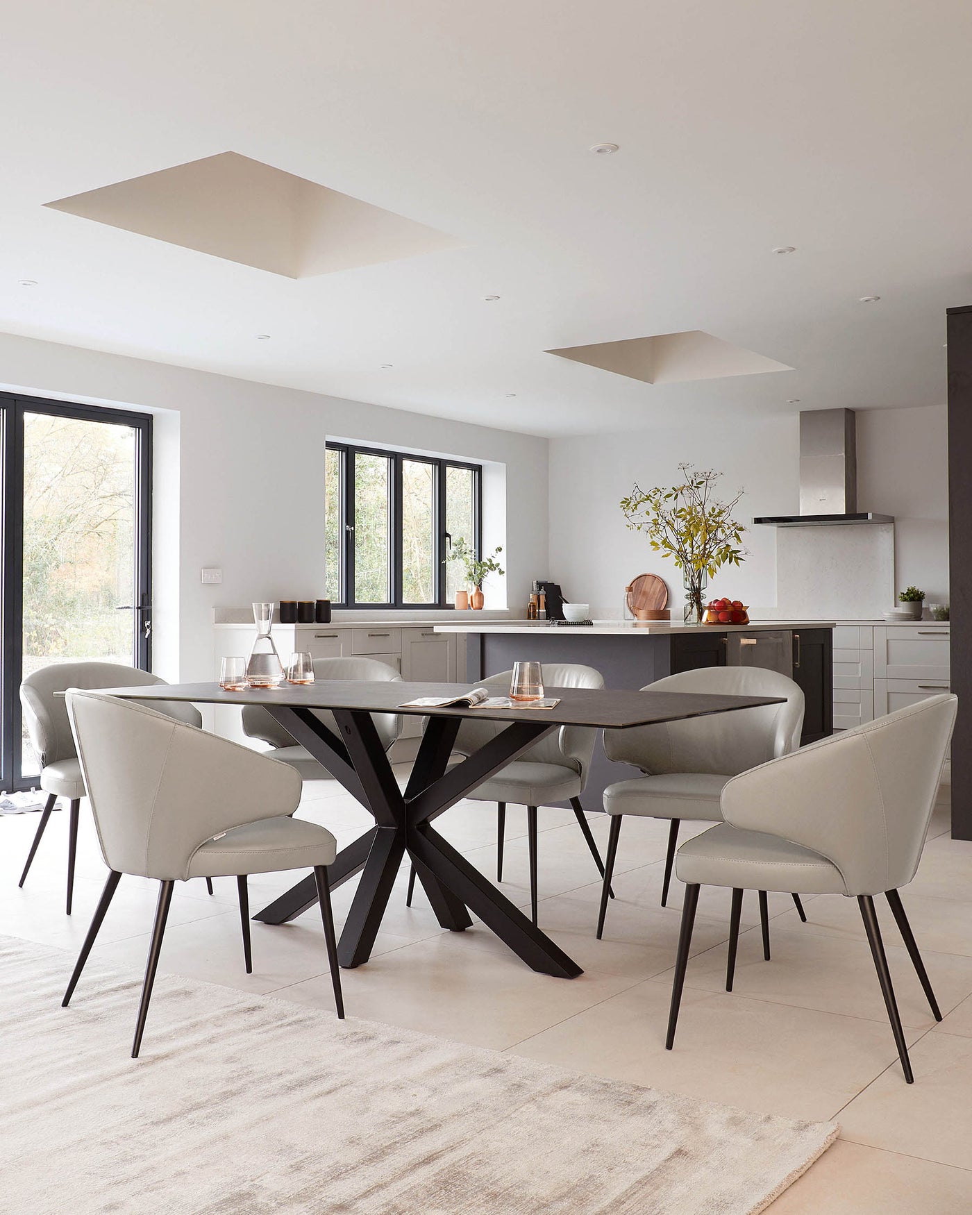 Modern dining room featuring a rectangular table with a unique, geometric black base and a smooth, dark tabletop. Surrounding the table are six elegant, upholstered chairs in light grey with slender black legs, designed for comfort and style. The setup is placed on a textured beige area rug, complementing the minimalist aesthetic of the room.