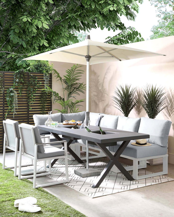 Modern outdoor dining set featuring a grey rectangular table with a unique X-shaped leg design, paired with six contemporary white-framed chairs with light grey seat and back cushions. A coordinating light grey outdoor sofa bench with plush cushions sits along one side of the table. The ensemble is complemented by a large off-white parasol, providing shade, all arranged on an elegant patterned outdoor rug, set against a backdrop of lush greenery and privacy screens.