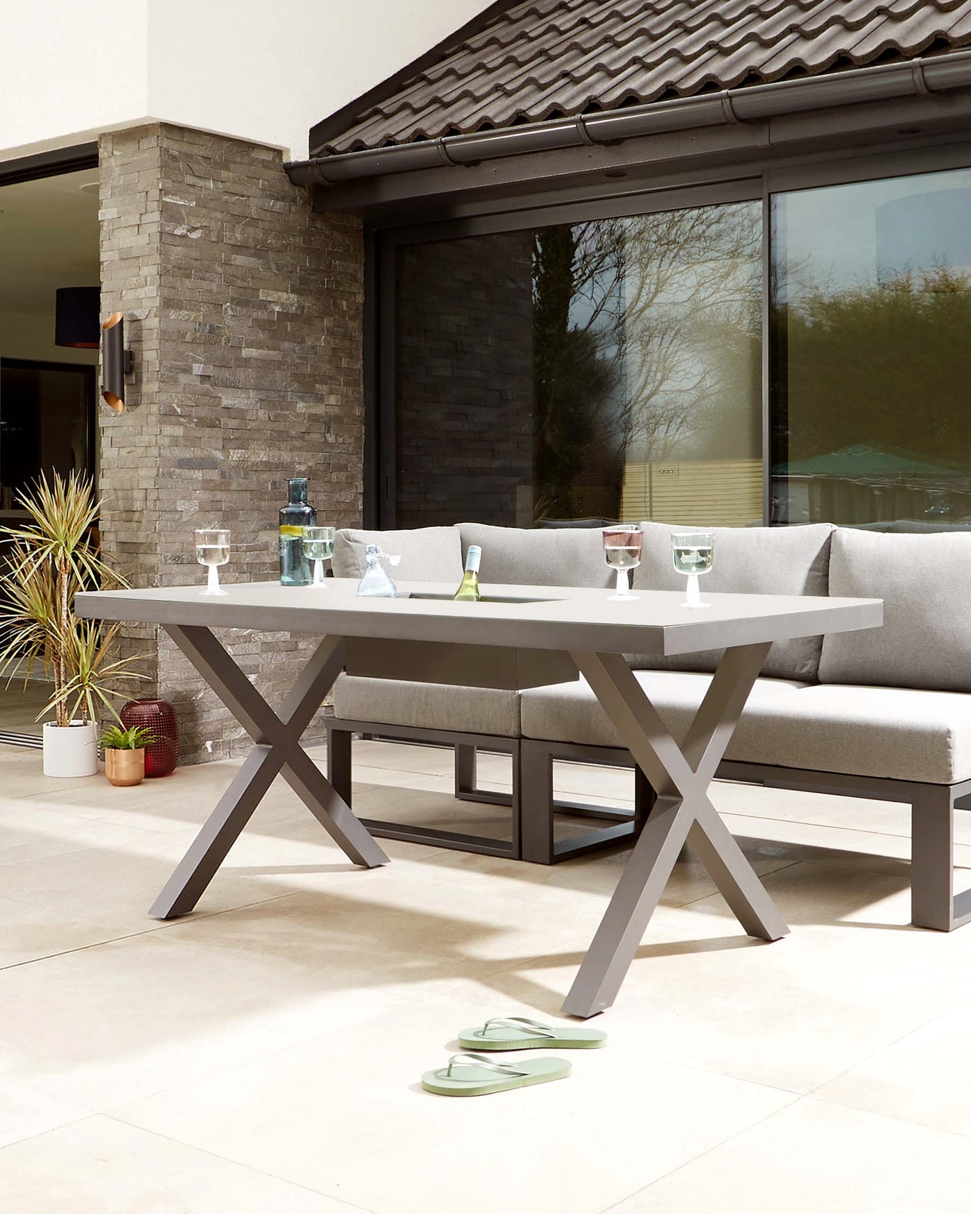 Contemporary outdoor furniture set featuring a grey rectangular dining table with a solid top and a distinctive X-shaped base, paired with a matching L-shaped sectional sofa with plush grey cushions, all arranged on a patio with a stone pillar and sliding glass doors in the background.