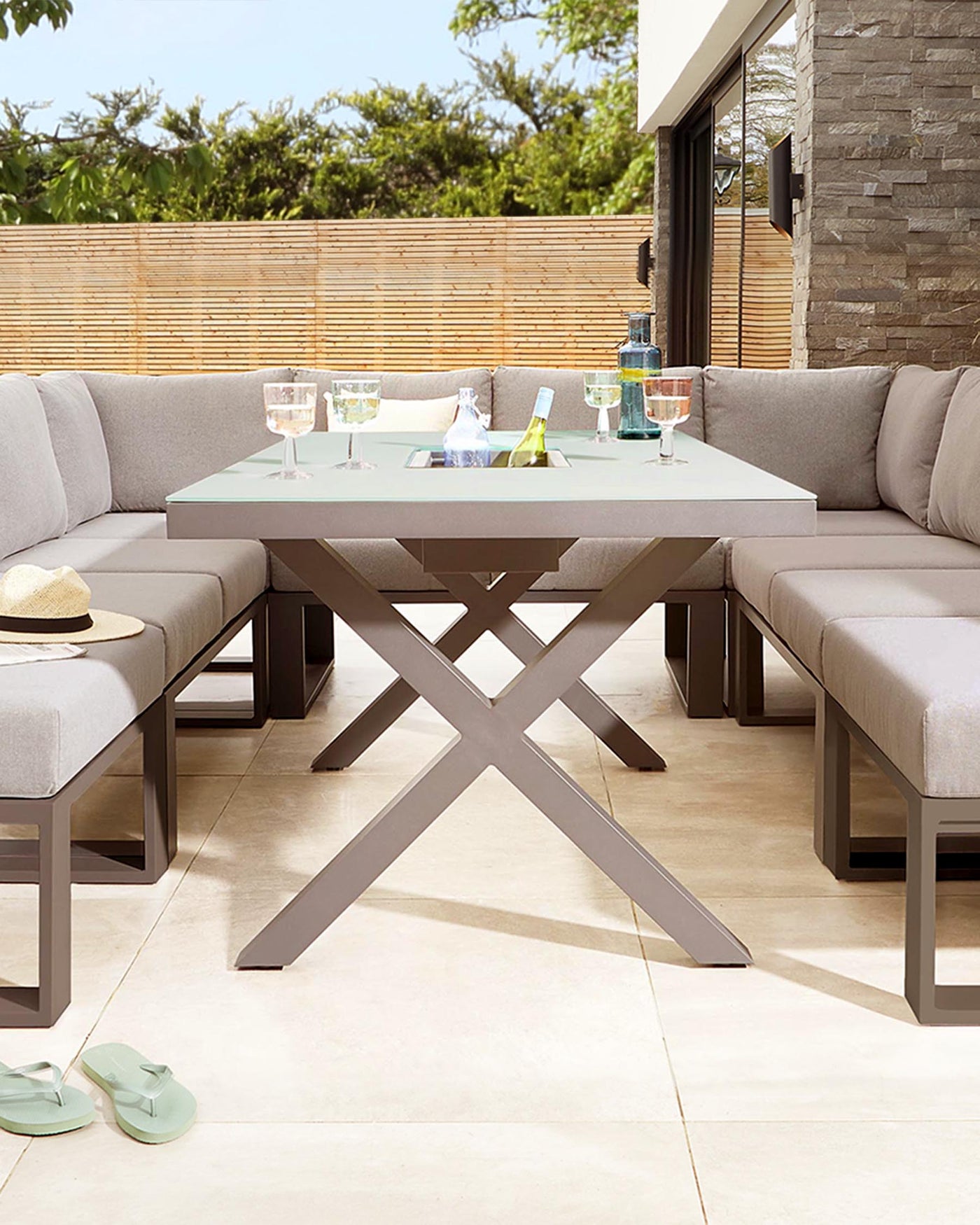 Outdoor patio furniture featuring a modern, rectangular dining table with a frosted glass top and crisscrossing metal legs in a taupe finish. Surrounding the table is an L-shaped sectional sofa with light grey cushions, providing comfortable seating for a relaxed outdoor dining experience. The neutral tones complement a contemporary design aesthetic.