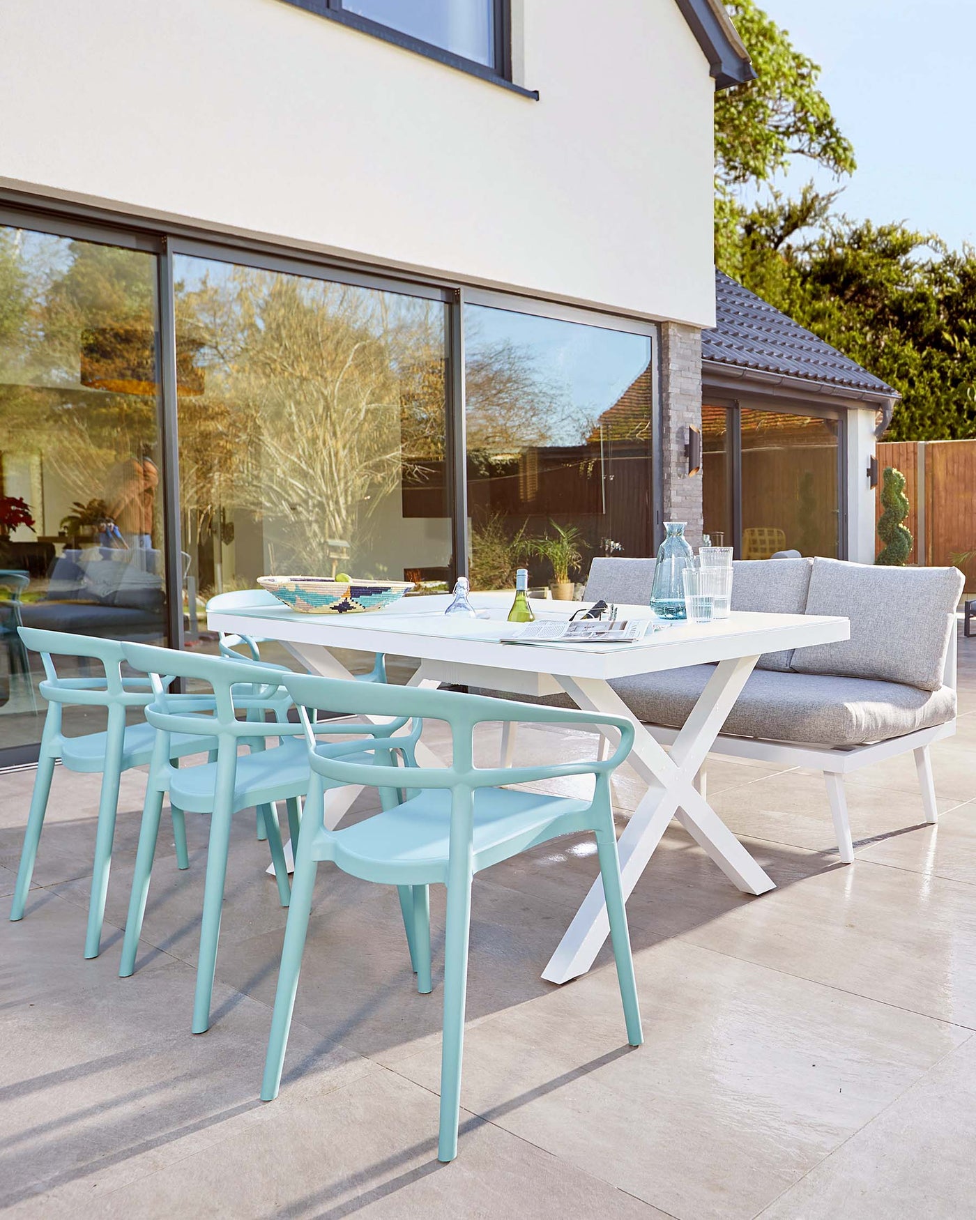 Modern outdoor dining furniture set featuring a white rectangular table with a clean design and cross-leg support, paired with six aqua blue, contemporary plastic chairs, and a comfortable lounge-style bench with grey upholstery on one side of the table. The set is on a patio area against a backdrop of sliding glass doors and a residential structure.