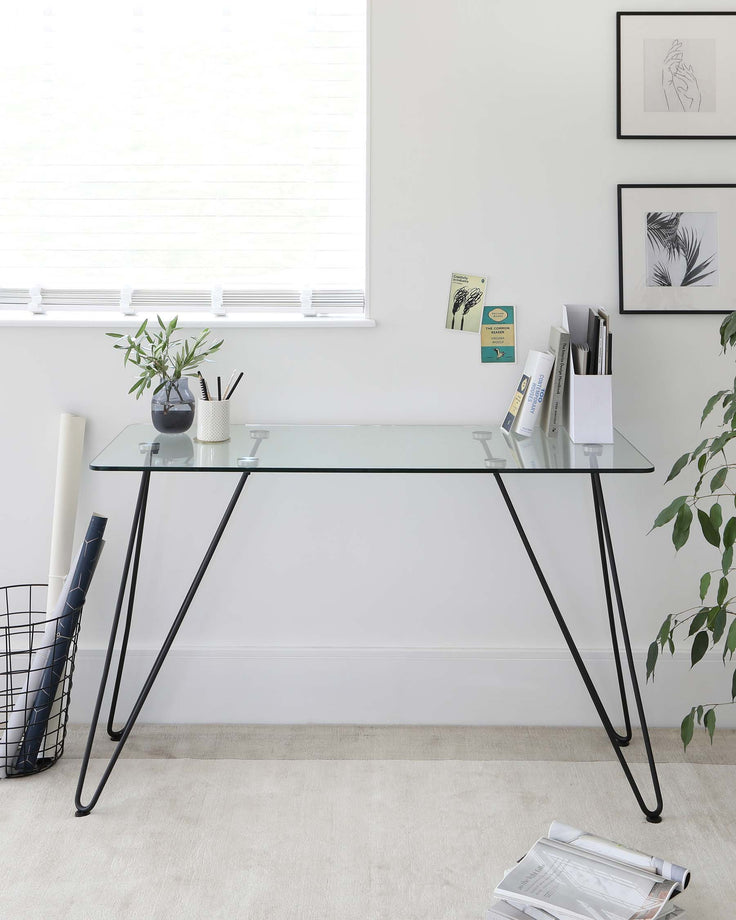 A minimalist modern desk with a clear glass tabletop and sleek black metal hairpin legs.