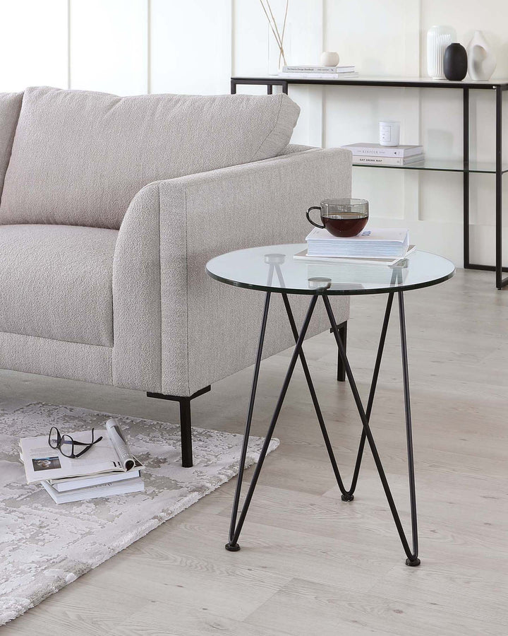 A modern, minimalist living room setup featuring a light grey textured fabric sofa with clean lines and a rounded armrest. Adjacent to the sofa is a round, glass-topped side table with a thin black metal frame forming a tripod support. On the table is a simple ceramic mug and a small stack of books. The setting includes a patterned off-white area rug, a sleek black metal shelving unit in the background holding decorative items, and a second stack of books and glasses on the floor beside the side table.