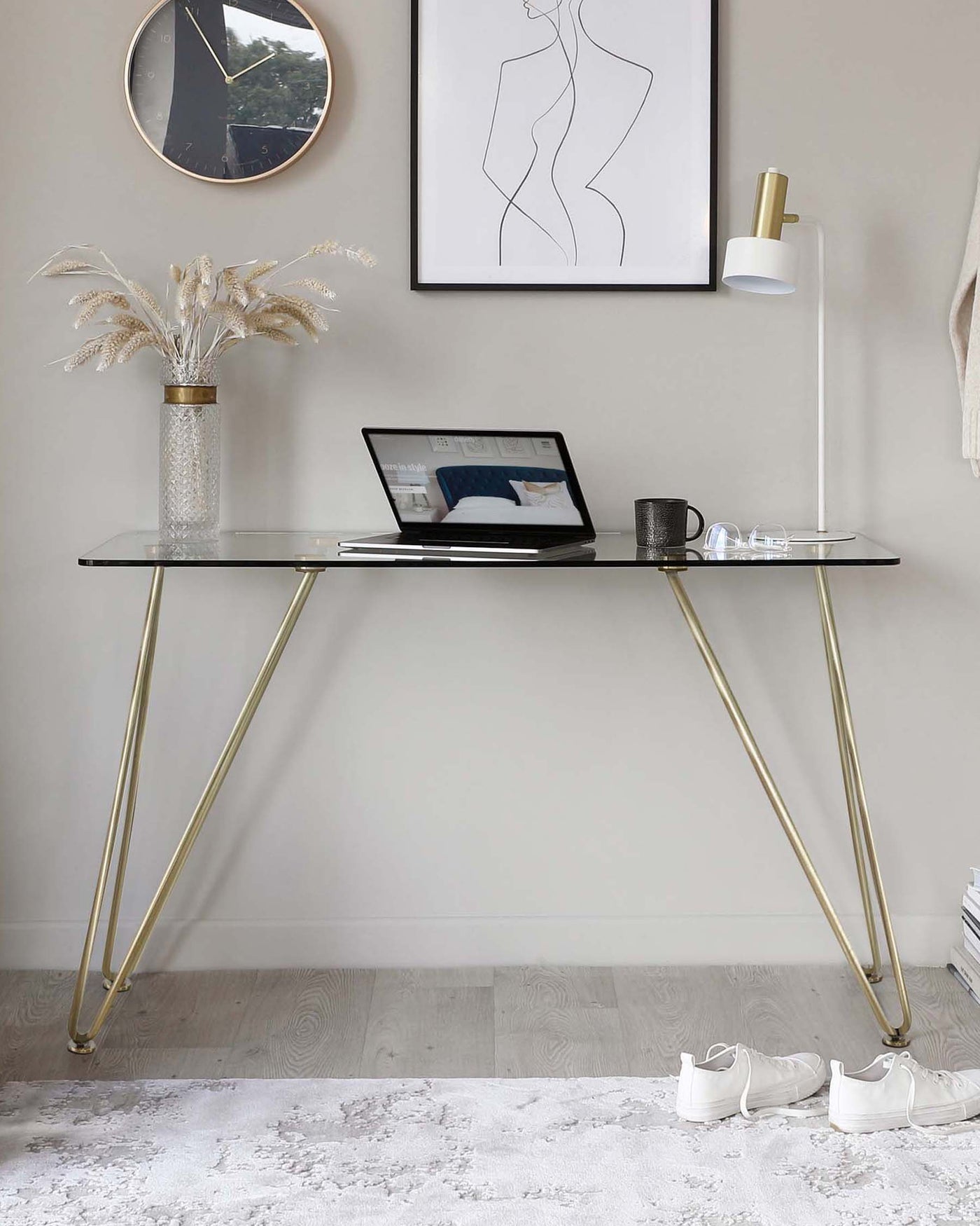Elegant and modern black glass top writing desk with sleek gold metal legs in a minimalist room setting, accompanied by a gold-and-white floor lamp, a decorative vase with dried plant arrangement, and framed artwork on the wall.