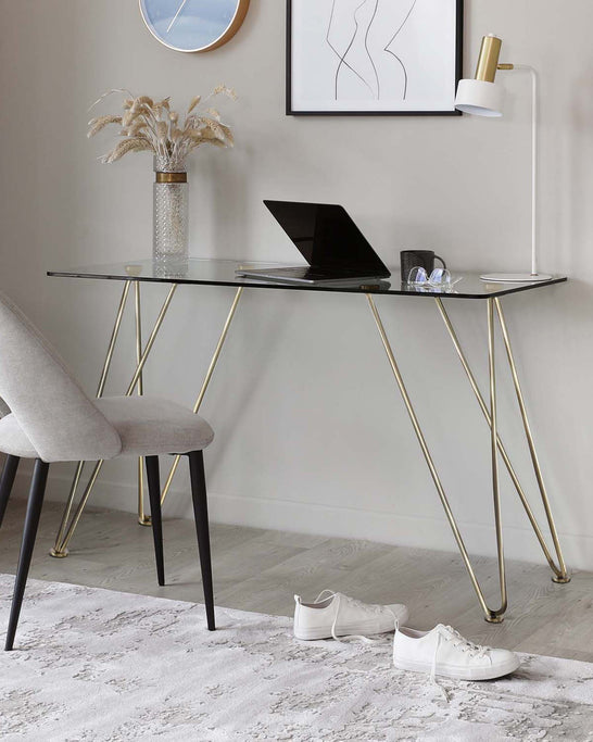 Elegant modern glass-top desk with gold-finished geometric metal legs. Coordinated with a minimalist light grey upholstered chair featuring sleek black wooden legs accented with gold tips.