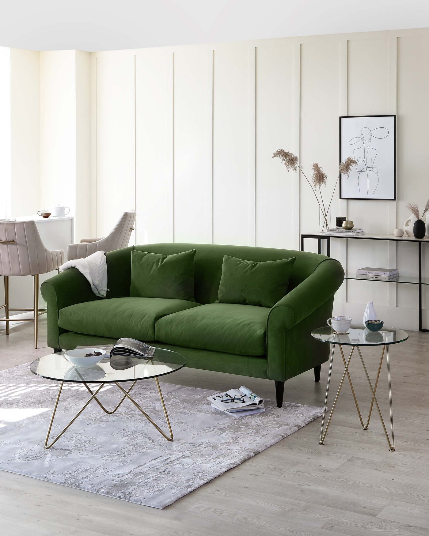 An elegant emerald green velvet sofa with a plush back and seating, accompanied by a pair of modern round glass-top side tables with golden metal legs. The room is further accented by a soft textured white area rug beneath the furniture.