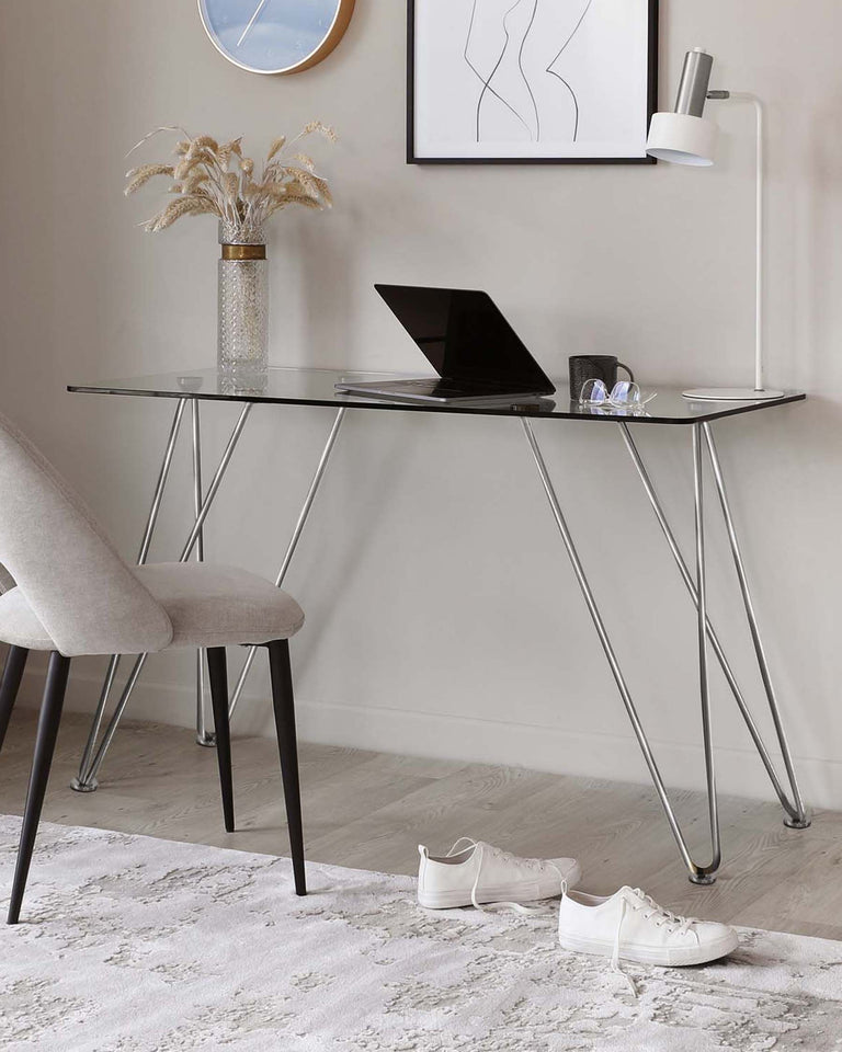 Modern minimalist furniture including a sleek, clear glass desk with crisscrossing silver metal legs and a comfortable-looking grey fabric upholstered chair with black tapered wooden legs. The classy and simple design of both pieces suggests versatility for various interior styles.