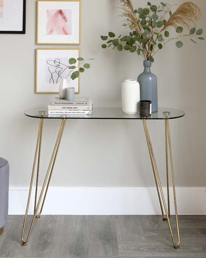 An elegant modern console table with a sleek, smoked glass top and slender, gently tapered, golden metal legs. The table is adorned with a curated selection of decorative items including books, a striated white vase, a muted blue bottle vase, a speckled black tumbler, and a small plant, and is positioned against a neutral wall with framed abstract art pieces above.