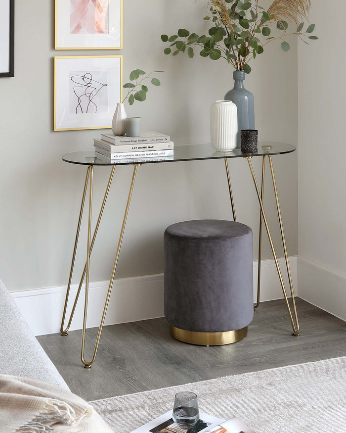 A modern oval console table with a glass top and slender golden metal legs, accompanied by a cylindrical grey velvet ottoman with a gold base. The tabletop is styled with decorative items including books, a vase, and a candle holder.