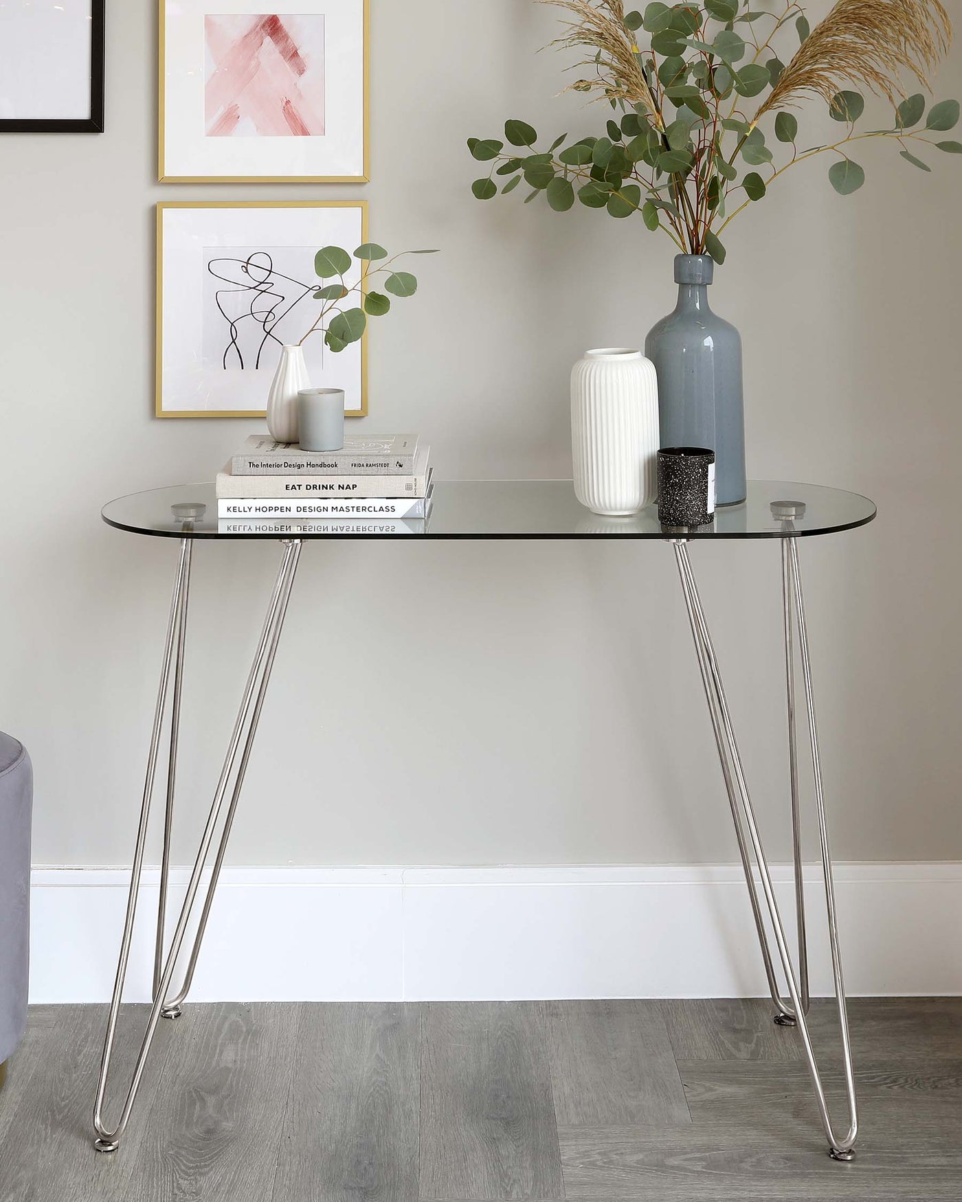 A modern, minimalist style glass console table with thin, tapered, high-shine chrome legs that cross under the tabletop forming a sleek, X-shaped base. The transparent glass top is rectangular with rounded edges, contributing to the table's elegant and contemporary design. The table is accessorized with decorative items, emphasizing its use as a stylish surface for displaying objects.