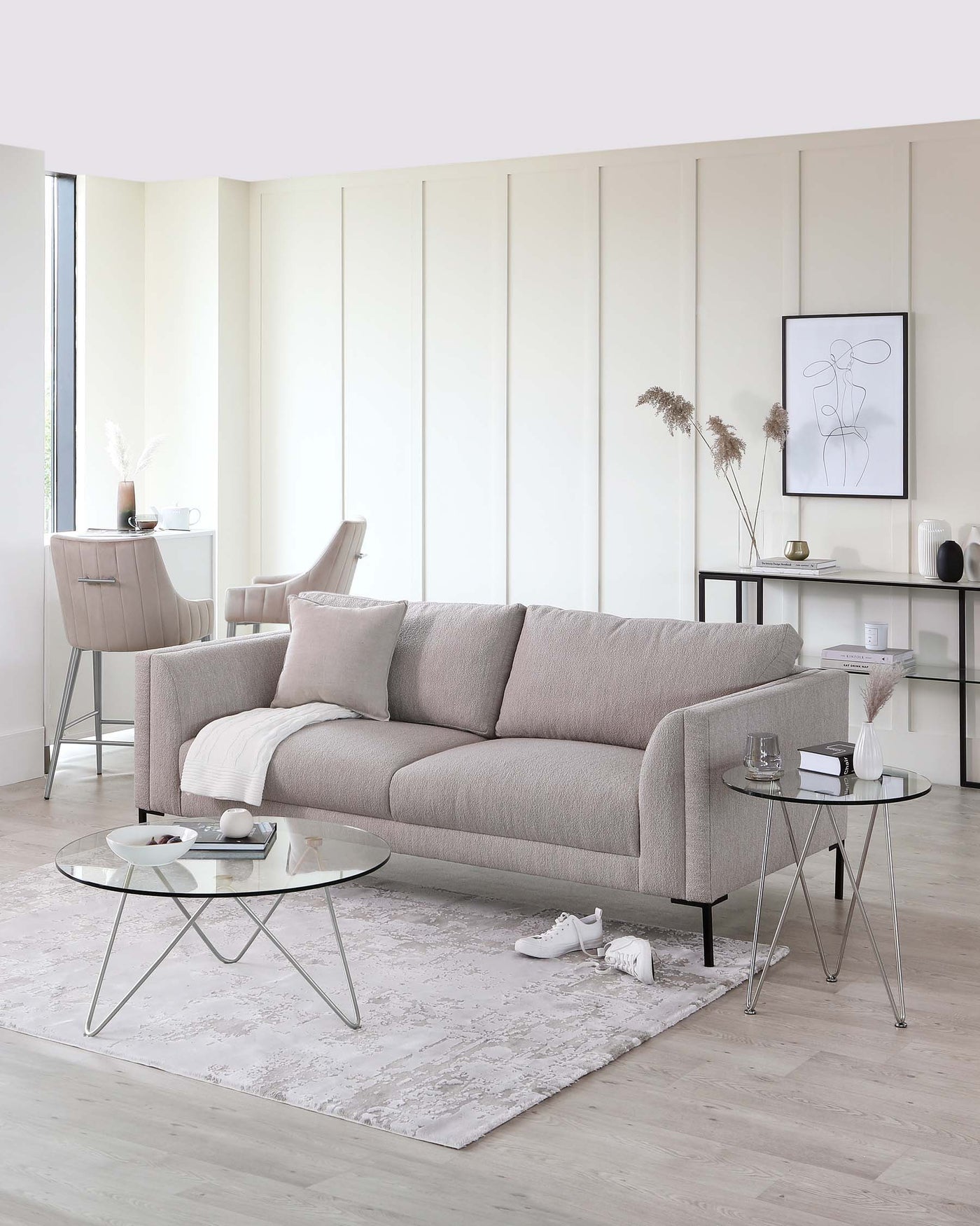 A contemporary living room setting featuring a three-seater fabric sofa in light grey with cushioned backrest and armrests. Accompanying the sofa is a round glass coffee table with a unique x-shaped metal base. Additionally, a matching side table with a glass top and slender metal legs is placed beside the sofa. The area is layered with a textured light grey area rug, adding warmth to the minimalist design.
