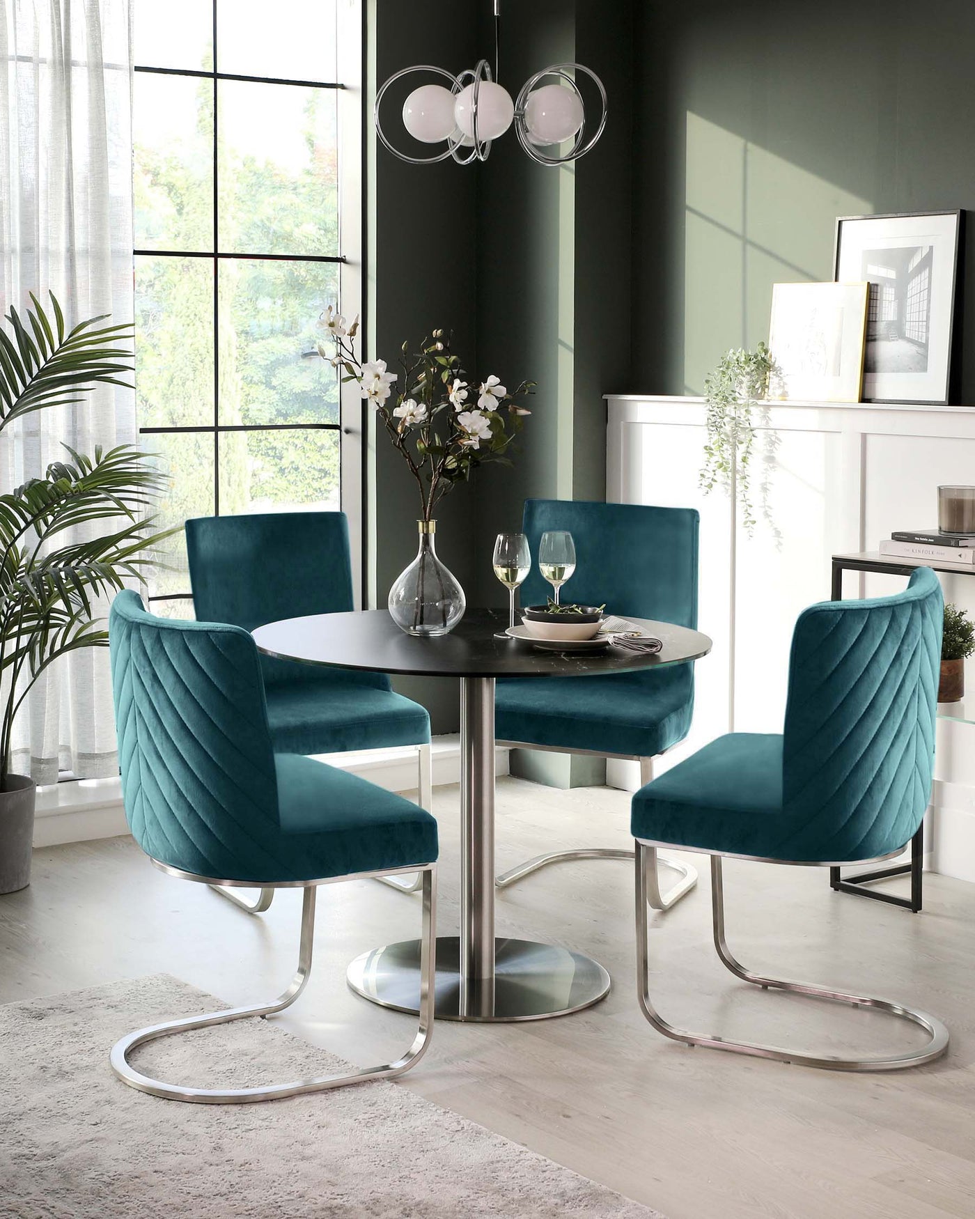 Modern dining room setup featuring an elegant round white table with a reflective metallic base and four luxurious teal velvet chairs designed with a unique quilted pattern and sleek silver cantilever frames.