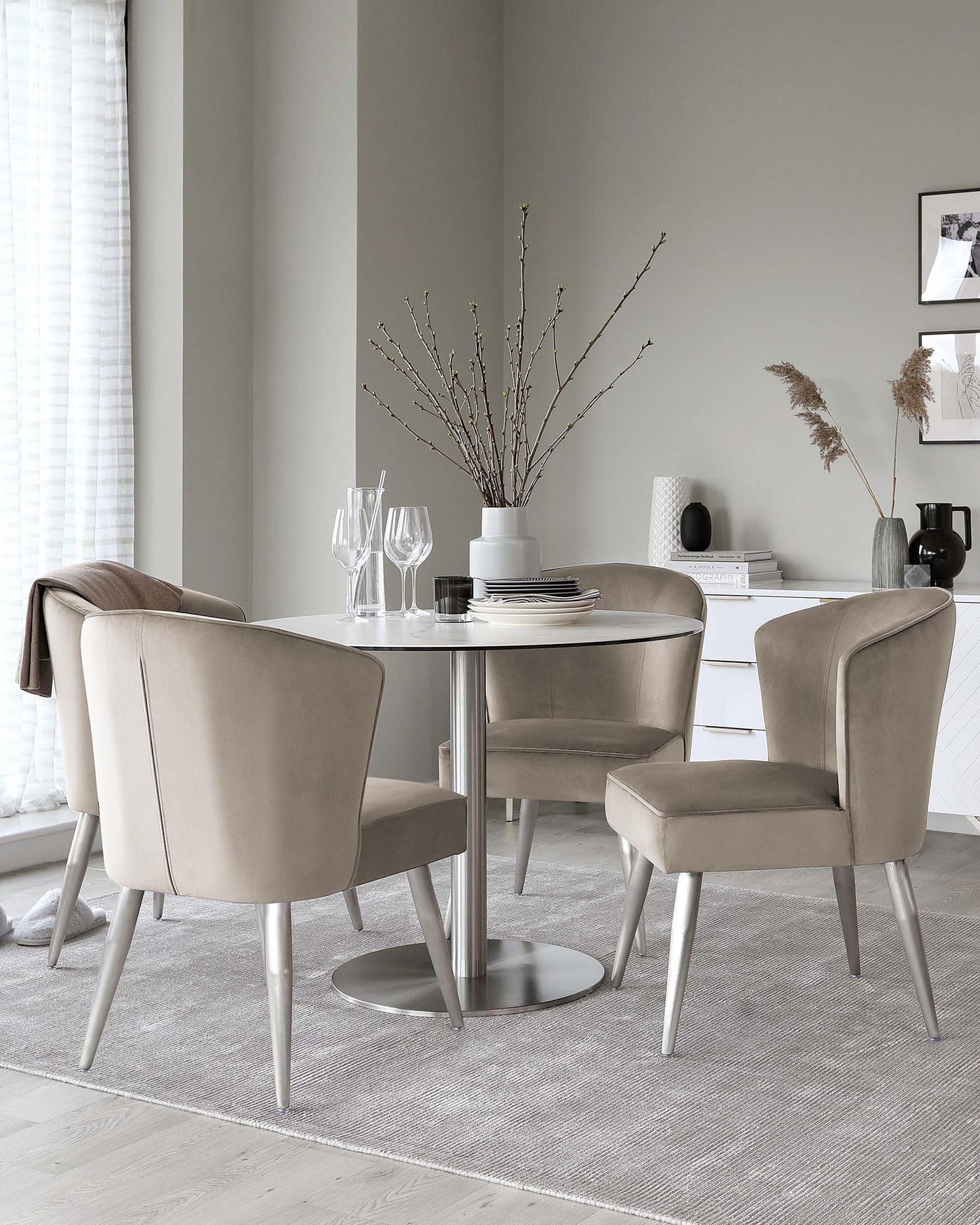 Round dining table with a reflective metal base and four matching upholstered chairs in a neutral tone on a textured grey rug.