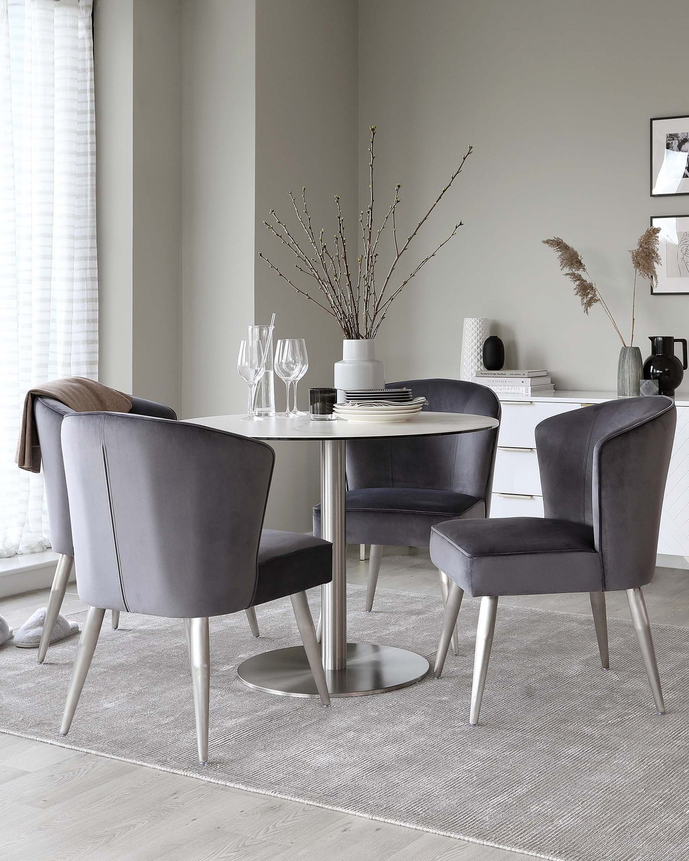 Modern dining room set with a round, chrome-legged table with a glossy finish and four plush, curved-back charcoal-grey chairs with tapered metal legs. The table is set with simple, elegant tableware and decorated with a vase of tall branches. A textured area rug underlies the setup, enhancing the sophisticated, contemporary feel of the space.