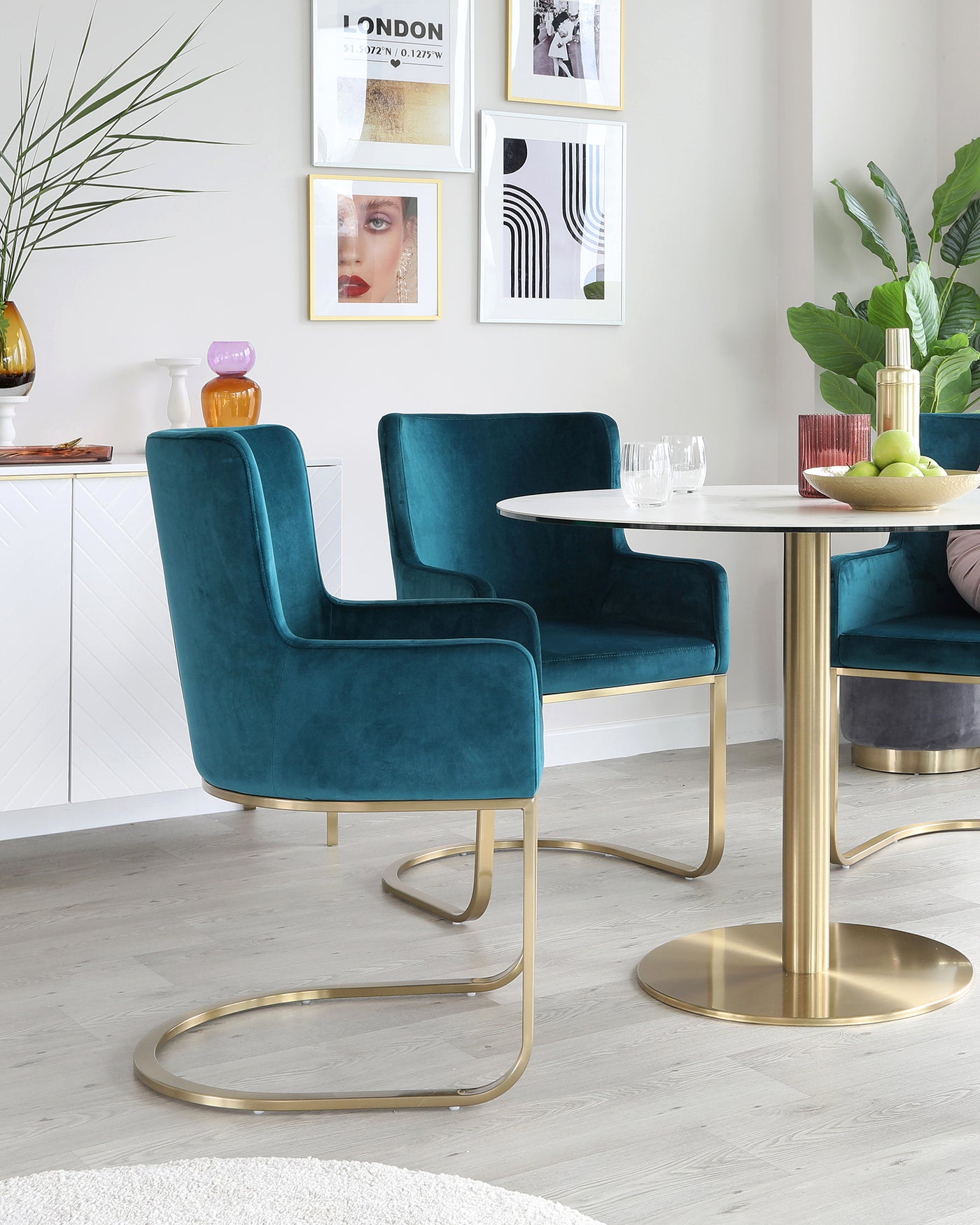 Two elegant teal velvet dining chairs with unique gold-finished metal legs, paired with a round white marble-top table with a gold-finished pedestal base.