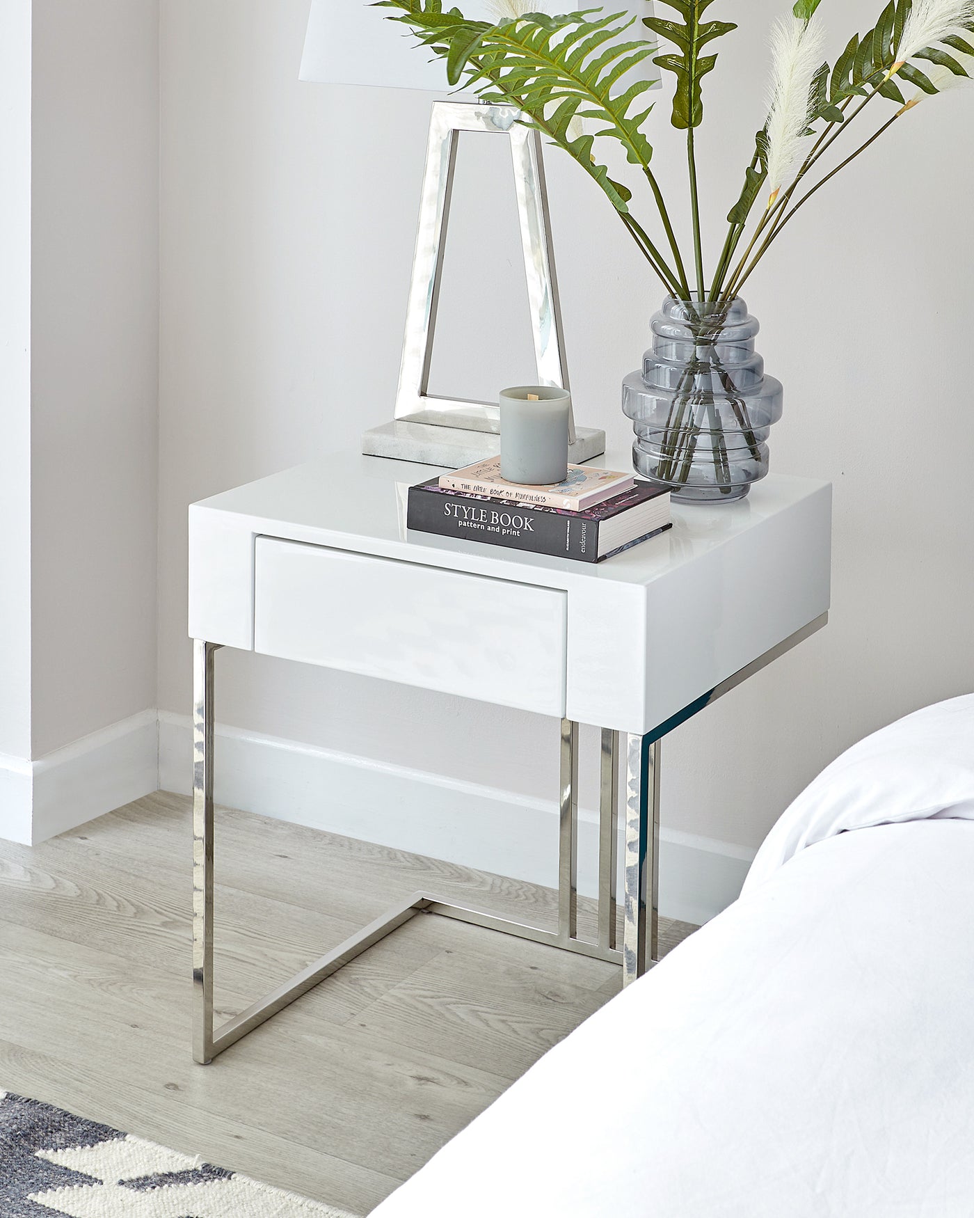 Modern minimalist white bedside table with a glossy finish and sleek silver metal legs, featuring a single drawer for storage.