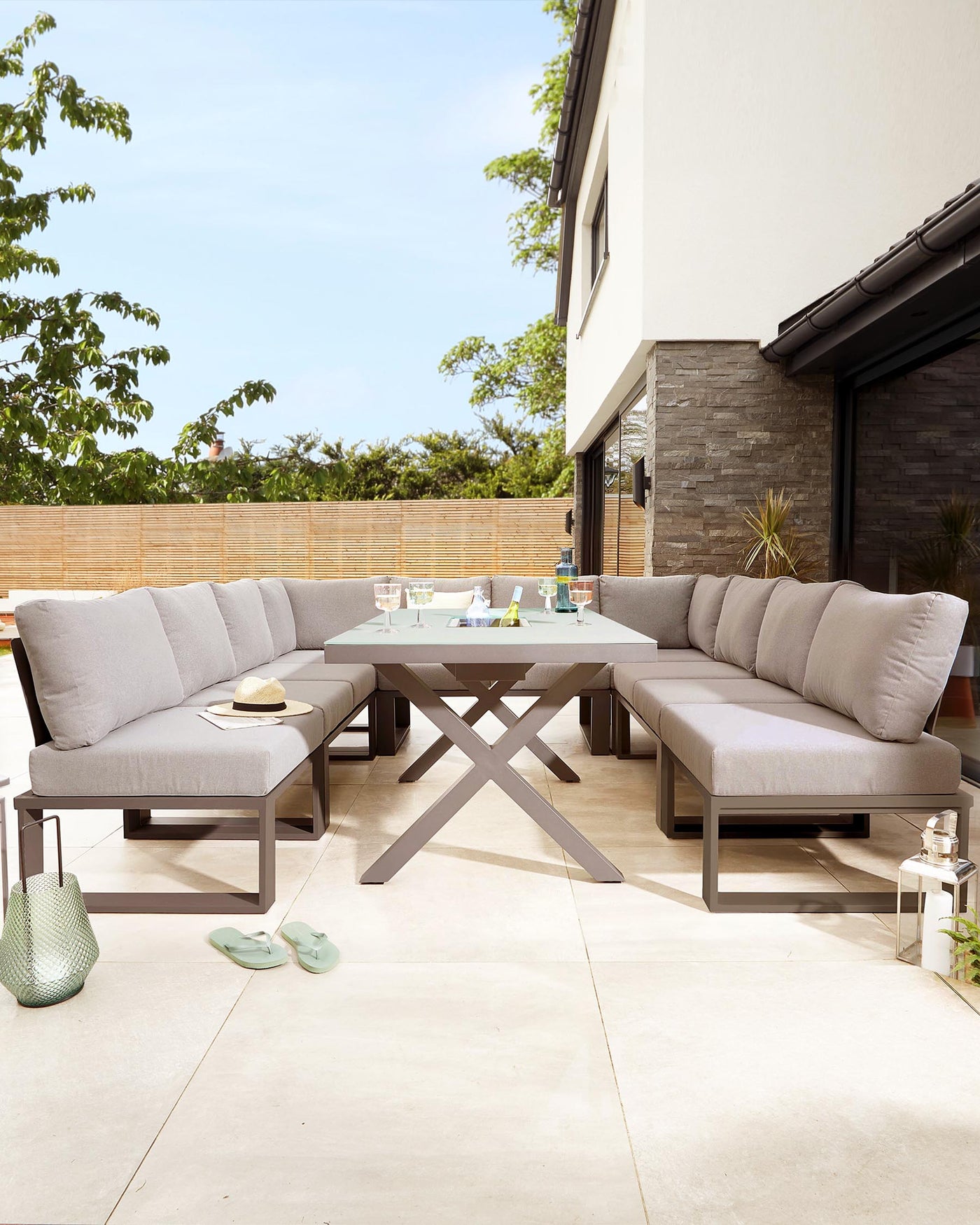 Outdoor furniture set featuring a modern rectangular dining table with a unique criss-cross leg design in a grey finish and a matching bench. The set also includes an L-shaped sectional sofa with plush cushions on a sleek grey frame, positioned on a neutral tiled patio, complemented by outdoor accessories.