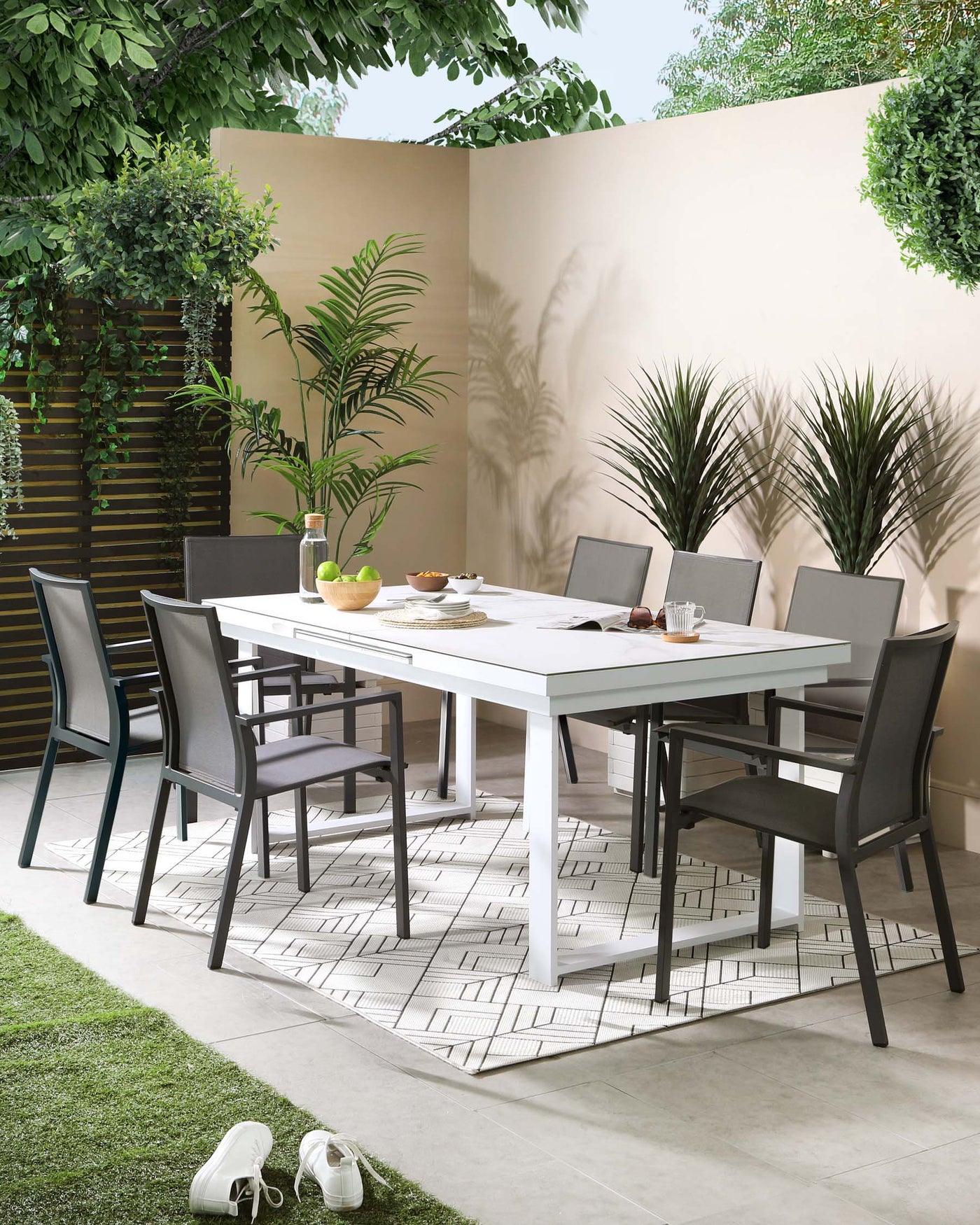 Modern outdoor dining set with a sleek white rectangular table and six charcoal grey chairs featuring clean lines and a minimalist design. The chairs have textile sling backrests and seats, providing both style and comfort for al fresco dining. The set is arranged on a geometric patterned area rug, complementing the contemporary aesthetic.