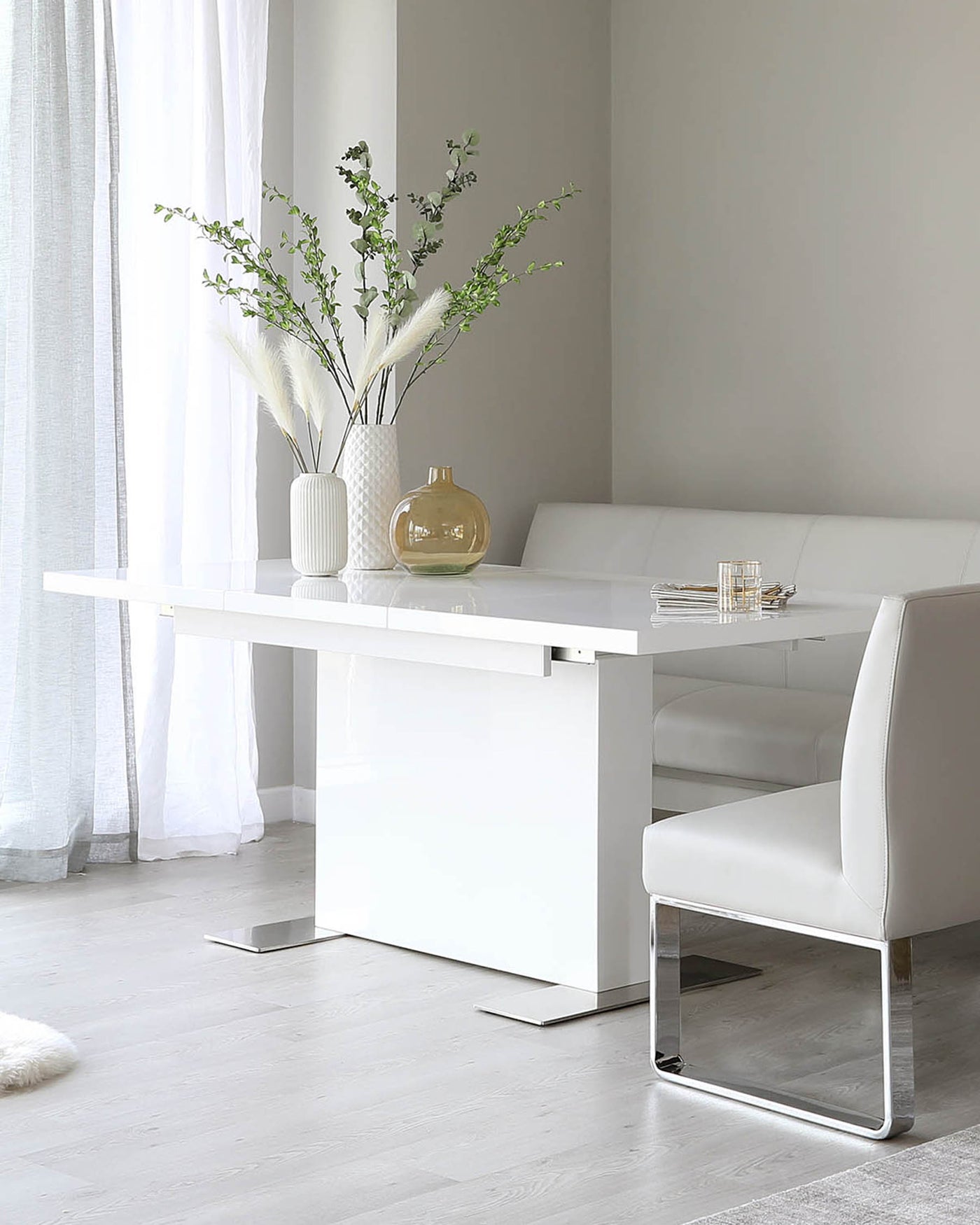 Modern minimalist dining set featuring a sleek white rectangular table with a pedestal base, paired with a matching white corner bench and a single white dining chair with chrome base. The set is complemented by elegant home decor including a vase with greenery, a decorative bottle, and a simple glass coaster set.