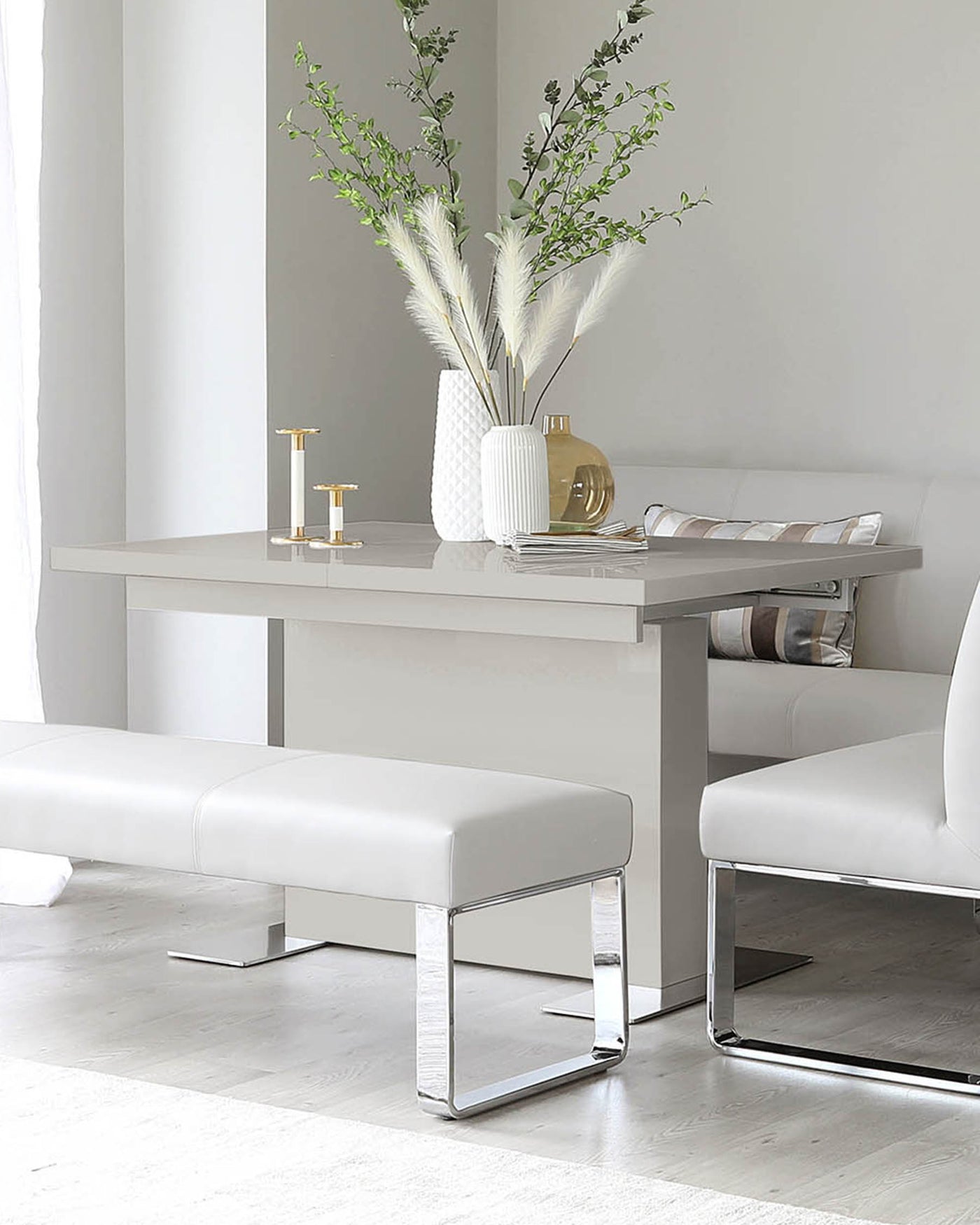Modern minimalist furniture in a neutral palette, featuring a sleek white rectangular dining table with a clean-lined design, complemented by two elegant white benches. Each bench has a padded seat for comfort and is supported by a striking chrome base that gives a contemporary touch to the ensemble. The scene is subtly decorated with a textured white vase containing pampas grass, golden decorative objects, and a stack of magazines, creating a sophisticated and inviting atmosphere.