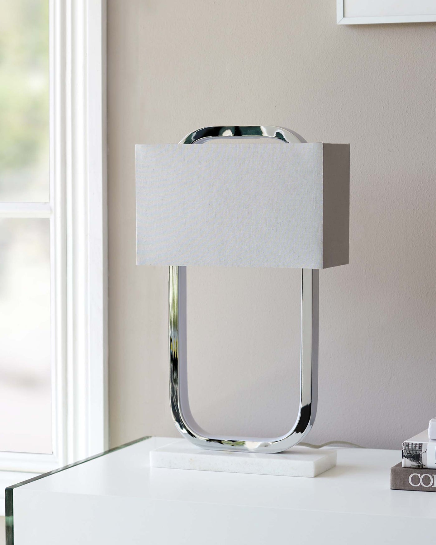 Modern table lamp with a chrome U-shaped base and white rectangular shade on a reflective white surface.