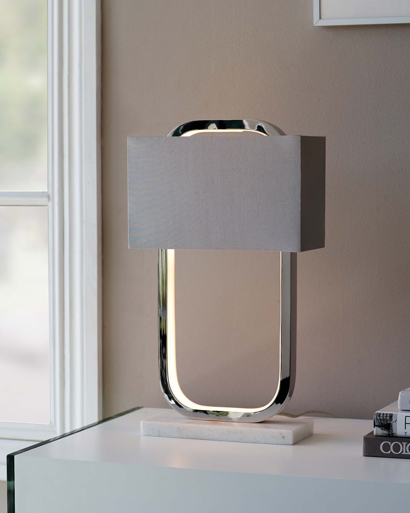 Contemporary table lamp with a polished chrome finish and an elegant fabric shade, placed on a glossy white surface near a window.