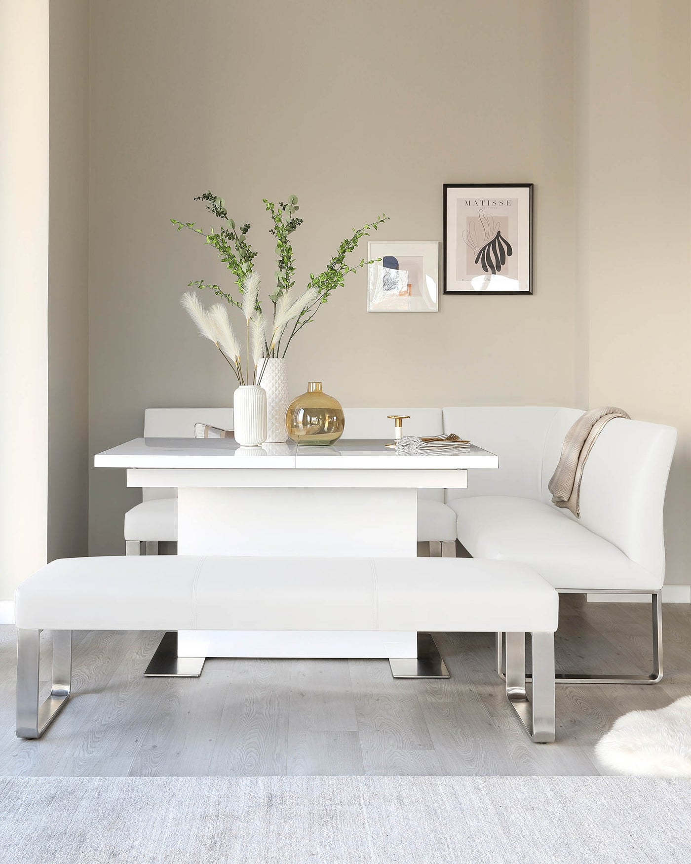 Modern minimalist white dining set including a rectangular table with a sleek surface and clean lines, accompanied by a matching bench and a curved-end L-shaped banquette, all with metallic silver legs.