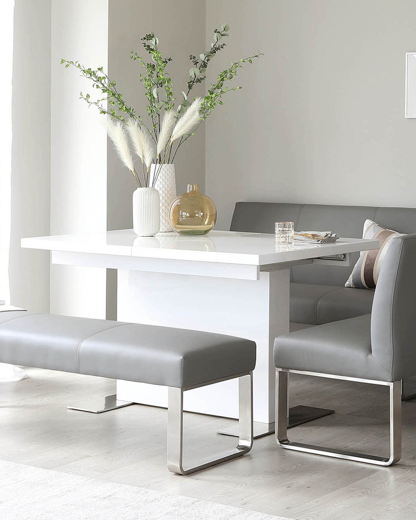 A modern dining set featuring a glossy white rectangular table with sleek, minimalistic lines and a set of two padded benches upholstered in light grey leatherette, each supported by lustrous chrome U-shaped legs. The ensemble suggests a contemporary, clean aesthetic for a sophisticated dining space.