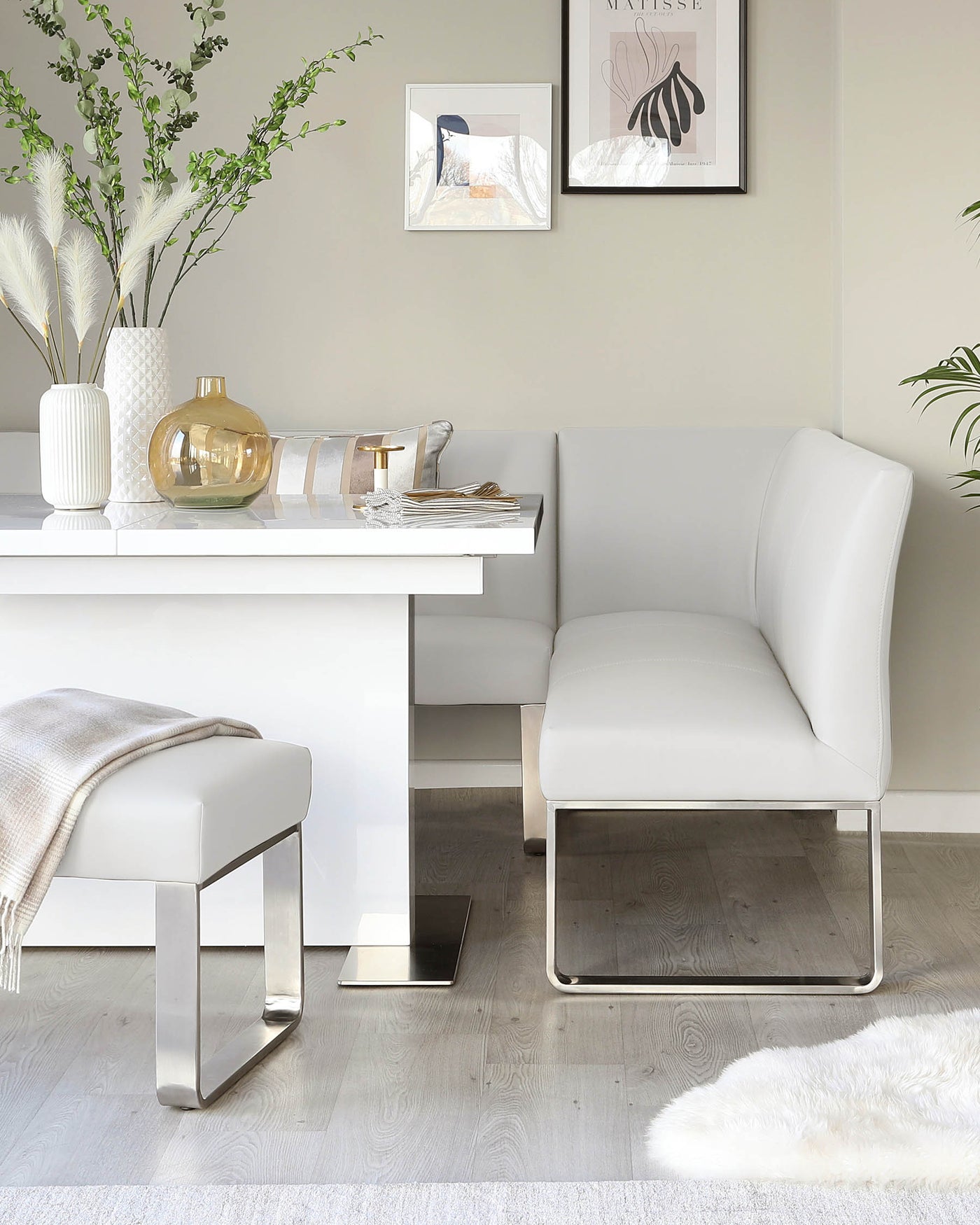 Modern minimalist furniture set featuring a sleek white dining bench with a high backrest, paired with a matching white rectangular dining table, both supported by clean-lined metallic bases. A simple yet elegant white ottoman with a metallic base complements the set, positioned beside the bench.