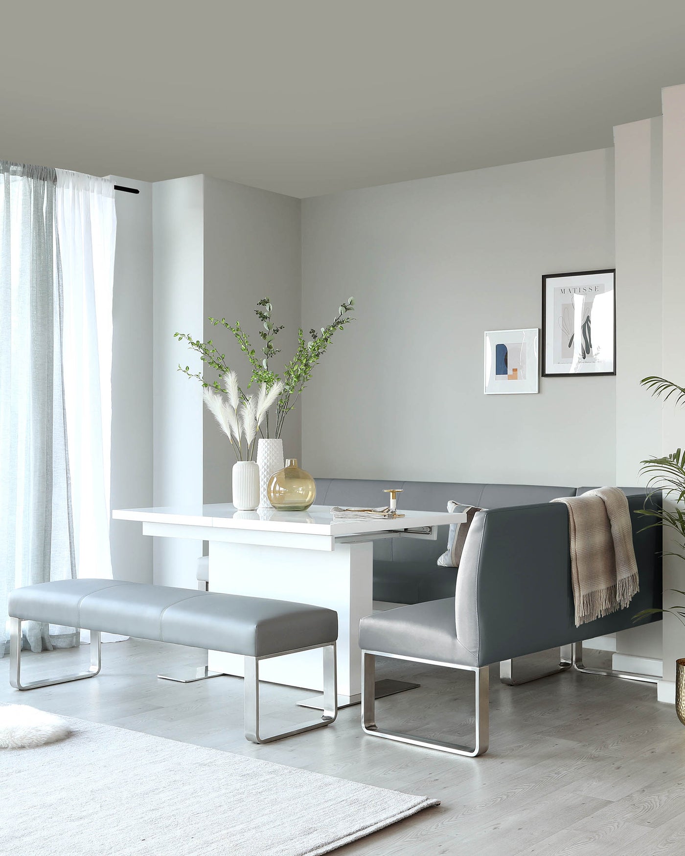 Modern minimalist furniture set featuring a sleek white rectangular dining table with a smooth finish, accompanied by a pair of grey upholstered benches with cushioned seats and chrome U-shaped legs, lending a contemporary and inviting aesthetic to the living space.