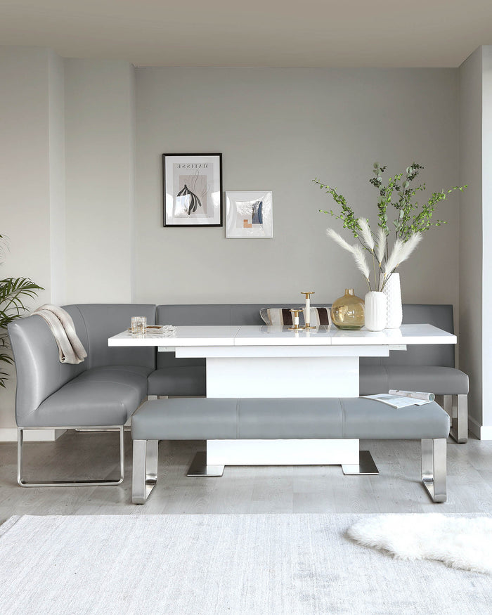 Modern minimalist furniture set in a dining space, featuring a sleek white rectangular table with a glossy finish, complemented by a grey upholstered L-shaped bench with clean lines and metallic chrome legs. The ensemble includes matching grey bench-style stools with a similar design aesthetic.
