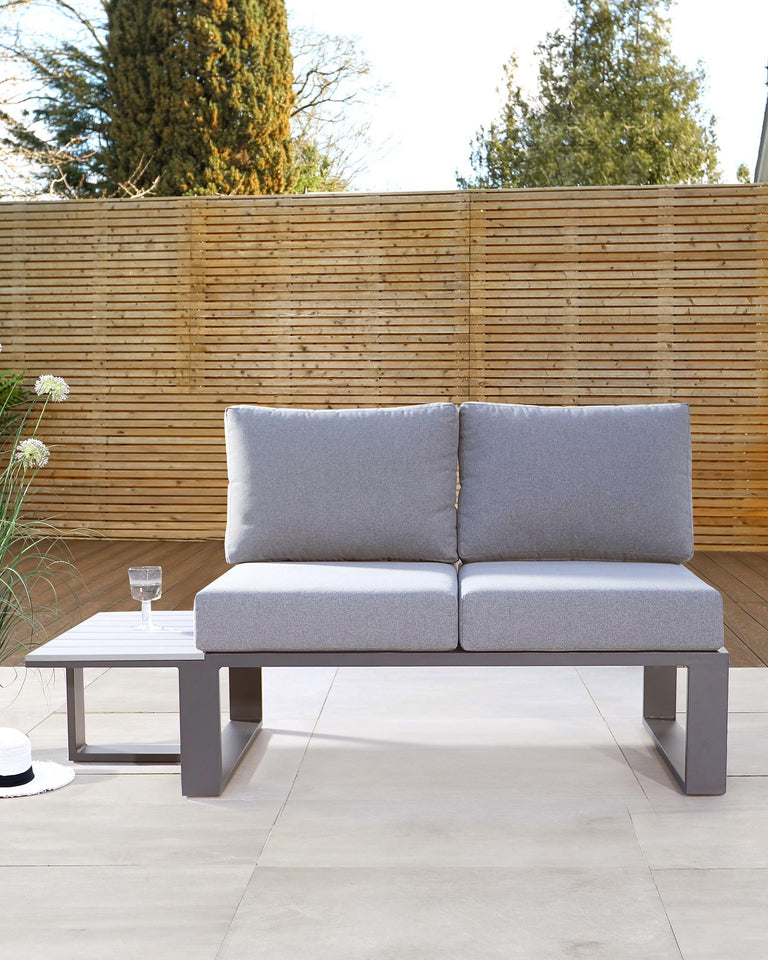 Modern two-seater outdoor sofa with grey cushions and a minimalist white square side table. The sofa features a sleek, metal frame with clean lines, and the side table complements the contemporary aesthetic. Both pieces are set against a bamboo fence backdrop, suggesting a tranquil garden setting.