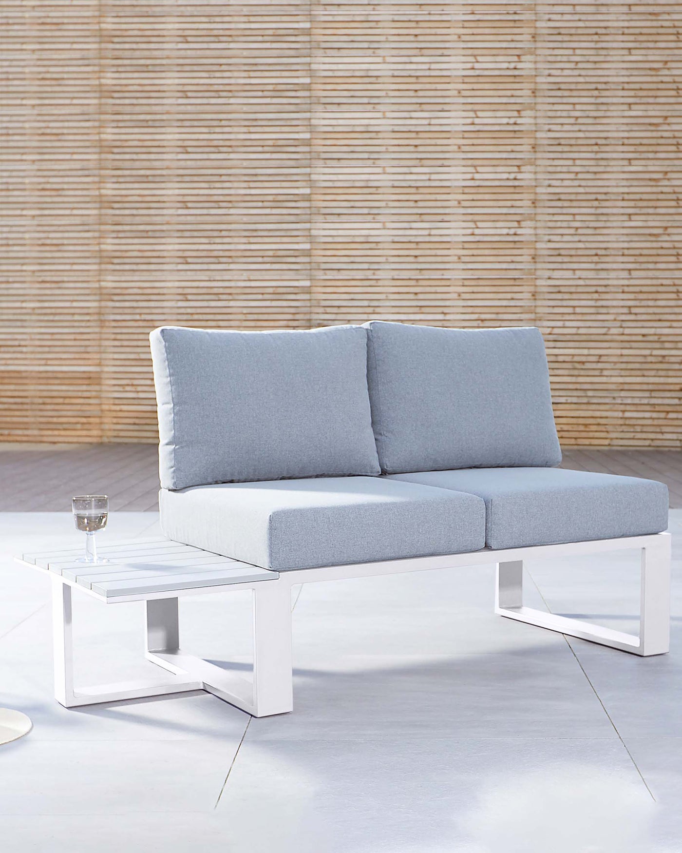 Modern two-seater outdoor sofa with light grey cushions and white metal frame, accompanied by a white rectangular low coffee table with a glass of water on top, positioned on a light concrete patio against a bamboo backdrop.