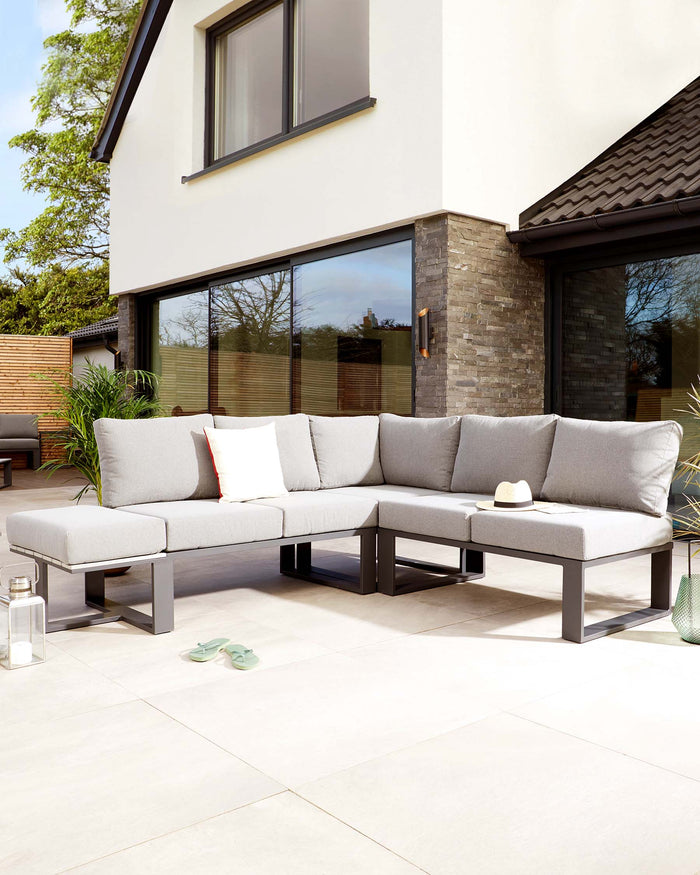 Modern outdoor L-shaped sectional sofa with light grey cushions on a sleek, dark metal frame, accompanied by a square metal ottoman, accessorized with white and orange throw pillows, displayed on a patio with neutral tiles.