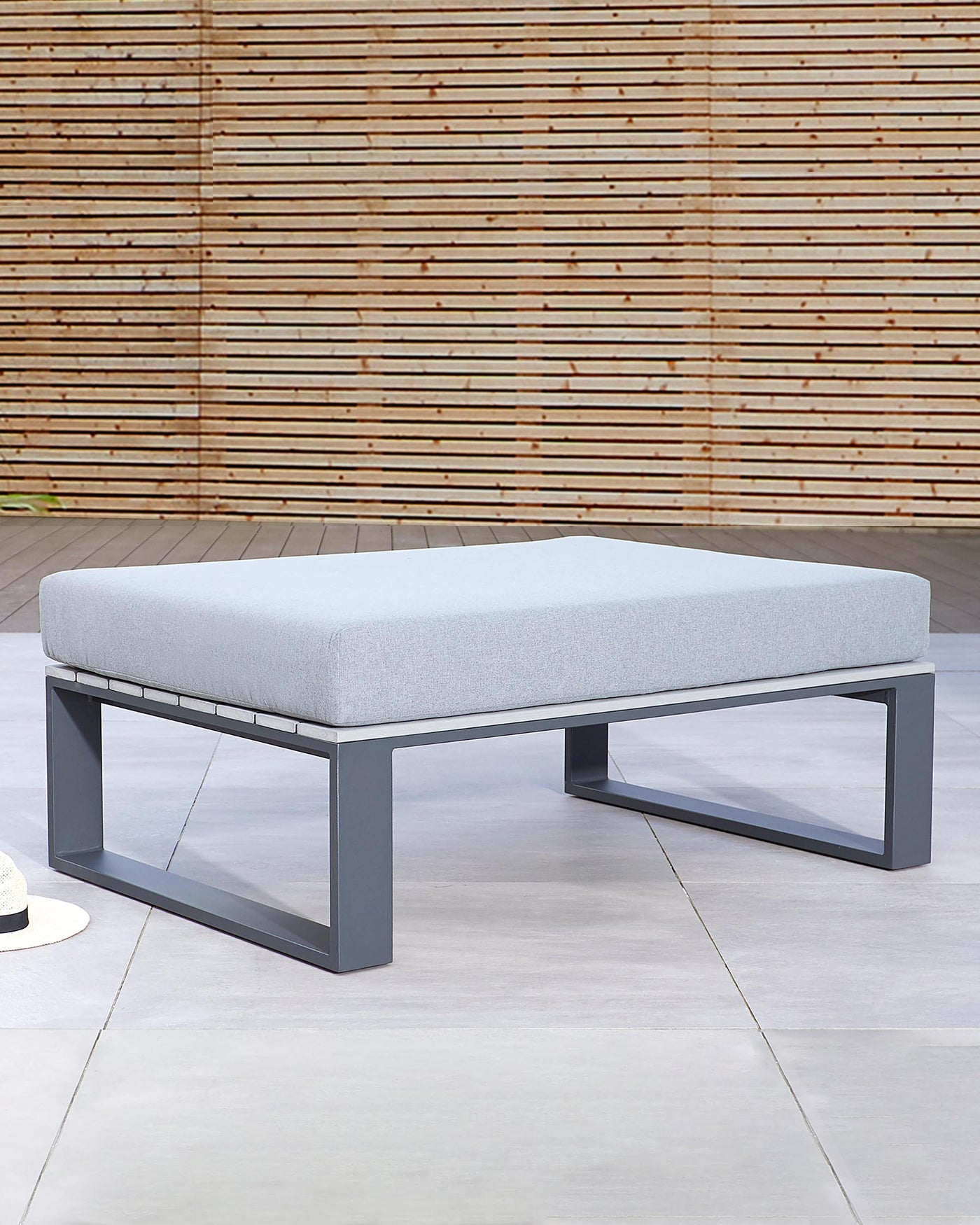 Modern rectangular ottoman with a light grey upholstered cushion top and a sleek, black metal base frame, set against a textured bamboo backdrop.