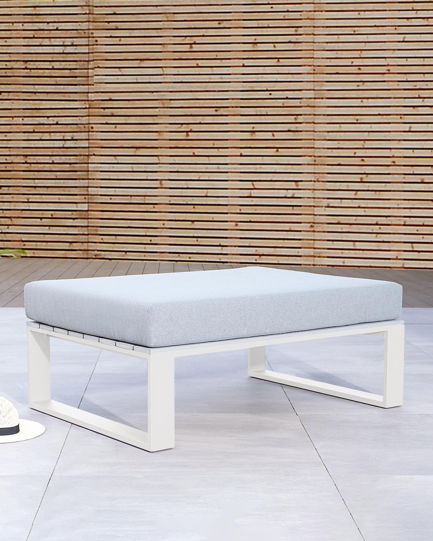 Modern outdoor ottoman with a white metal frame and light grey cushion, displayed on a pale floor against a wooden slat backdrop.