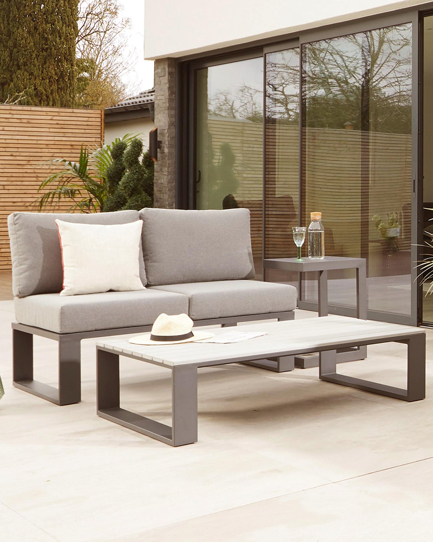 Modern outdoor furniture set featuring a grey, two-seater sofa with clean lines and plush cushions, accompanied by a low-profile, rectangular coffee table with a sleek grey finish. A matching side table with a clear glass top completes the set. All pieces have a contemporary metal frame design, suitable for elegant patio or garden spaces.