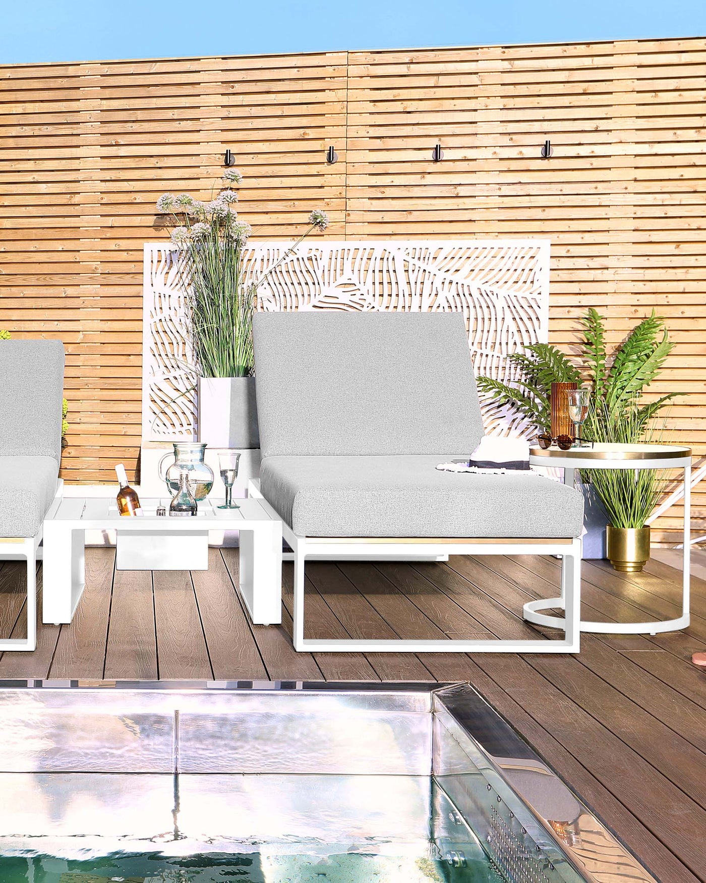 Modern outdoor furniture set on a wooden deck featuring a white-framed sofa with light grey cushions, a matching armchair, a white square coffee table, and a round side table with a golden base. The setting is accessorized with plants, decorative items, and is adjacent to a reflective pool.