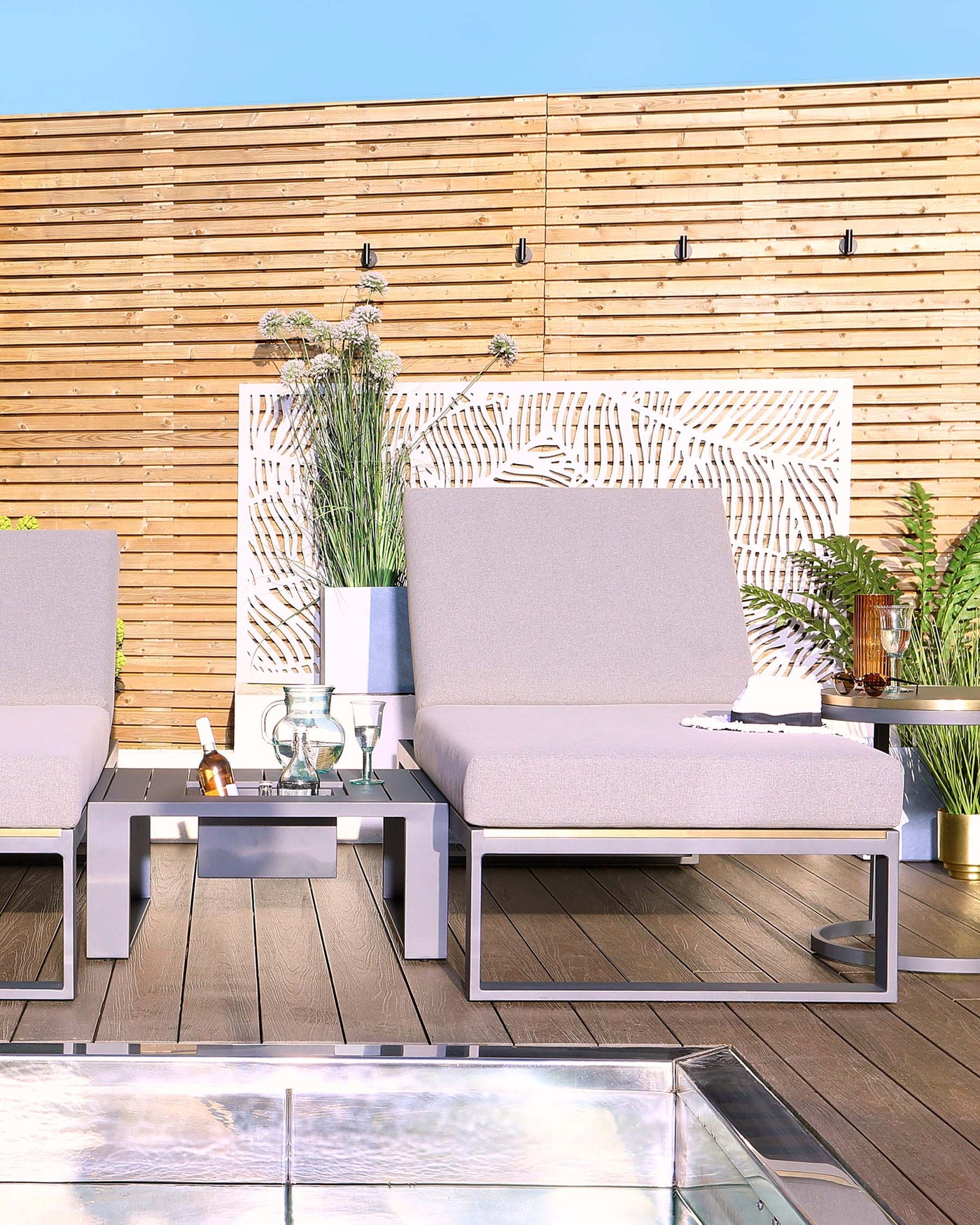 Modern outdoor patio furniture set featuring two sleek grey upholstered armchairs with white metal frames, complemented by a matching low-profile coffee table with a rectangular glass top. The set is arranged on a wooden deck, enhancing a contemporary and inviting outdoor space.