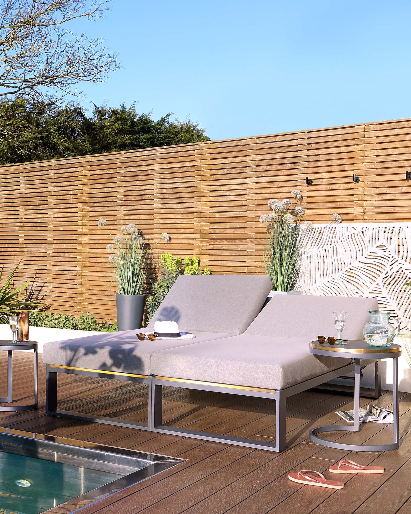 Modern outdoor furniture set featuring a sleek double lounger with light grey cushions and metal frame, complemented by a pair of cylindrical side tables with metal bases and wood-finish tops, all arranged on a wooden deck by a poolside.