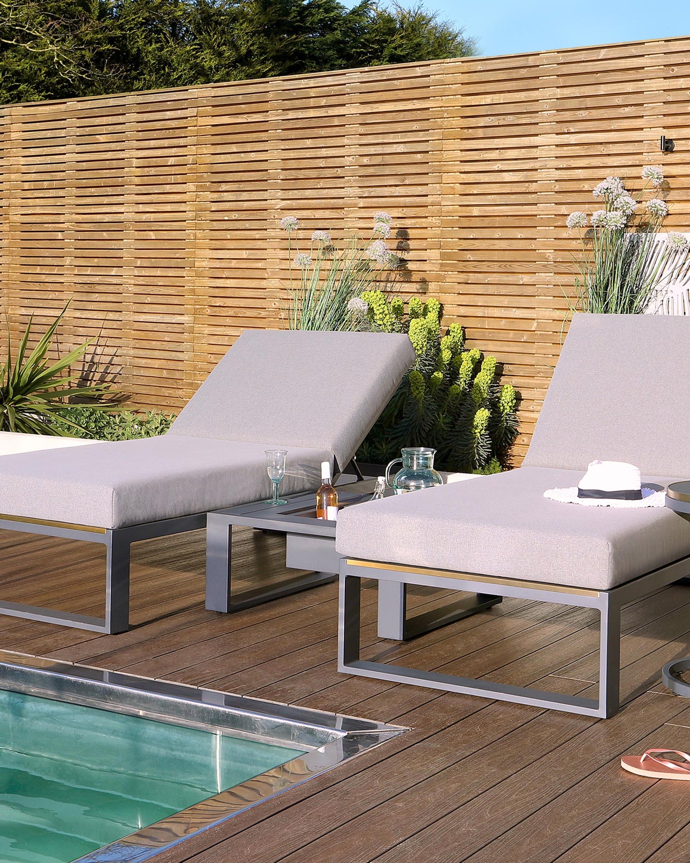 Two modern outdoor lounge chairs with light grey cushions and sleek grey metal frames, situated next to a small matching metal side table with a glass top, all displayed on a wooden deck beside a pool, complemented by a background of lush plants and a wooden slat fence.