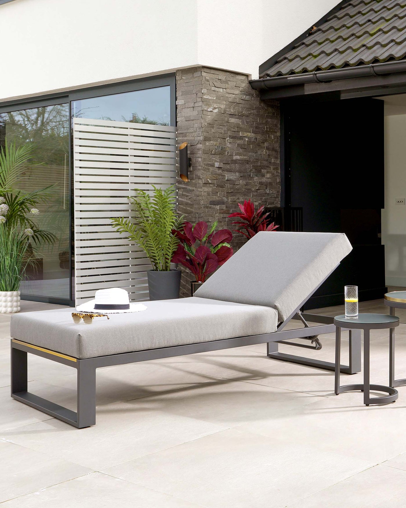 Modern outdoor furniture set featuring a sleek, adjustable chaise lounge with a matte black frame and light grey cushion, accompanied by a round, minimalistic matte black side table. The set is staged on a smooth patio surface with a contemporary backdrop of a home exterior, highlighting the furniture's suitability for upscale outdoor living spaces.