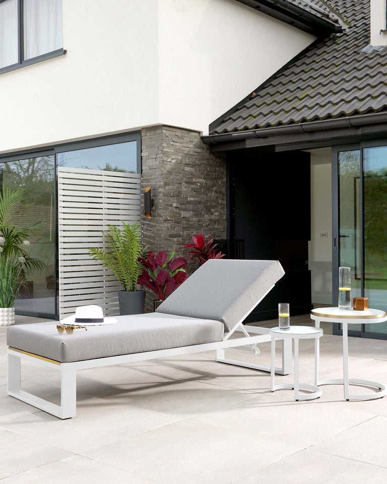 Modern outdoor furniture set featuring a minimalist white metal frame chaise lounge with a light grey cushion and an adjustable backrest, paired with a small round white side table with a two-tiered design. The set is displayed on a patio area outside a contemporary house, emphasizing sleek design and comfort.