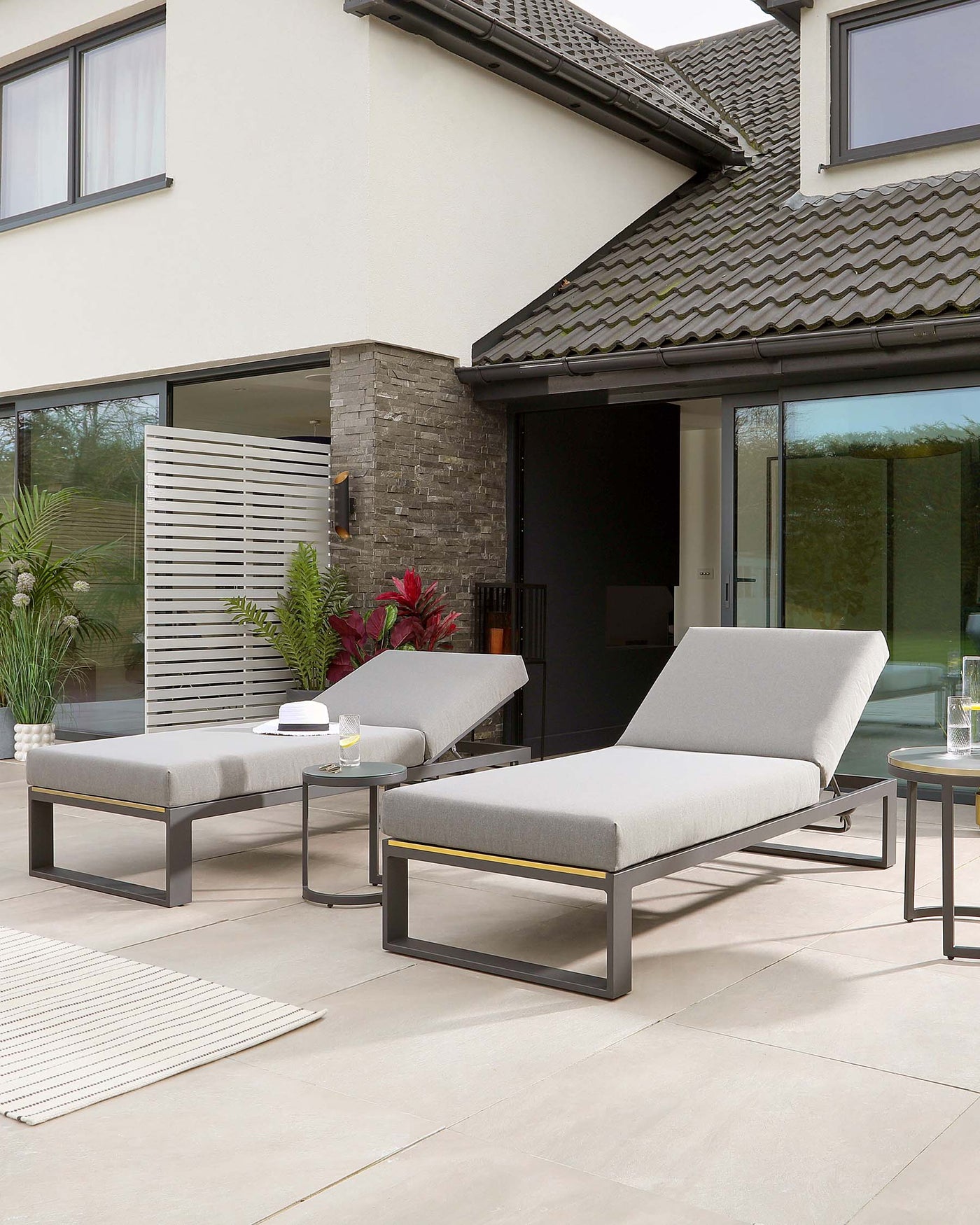 Modern outdoor furniture set featuring a pair of sleek, grey cushioned chaise lounges with matching black metal frames, accentuated with elegant gold detailing. A low, rectangular side table with a black metal frame and a glass top completes the ensemble, arranged on a paved patio area.