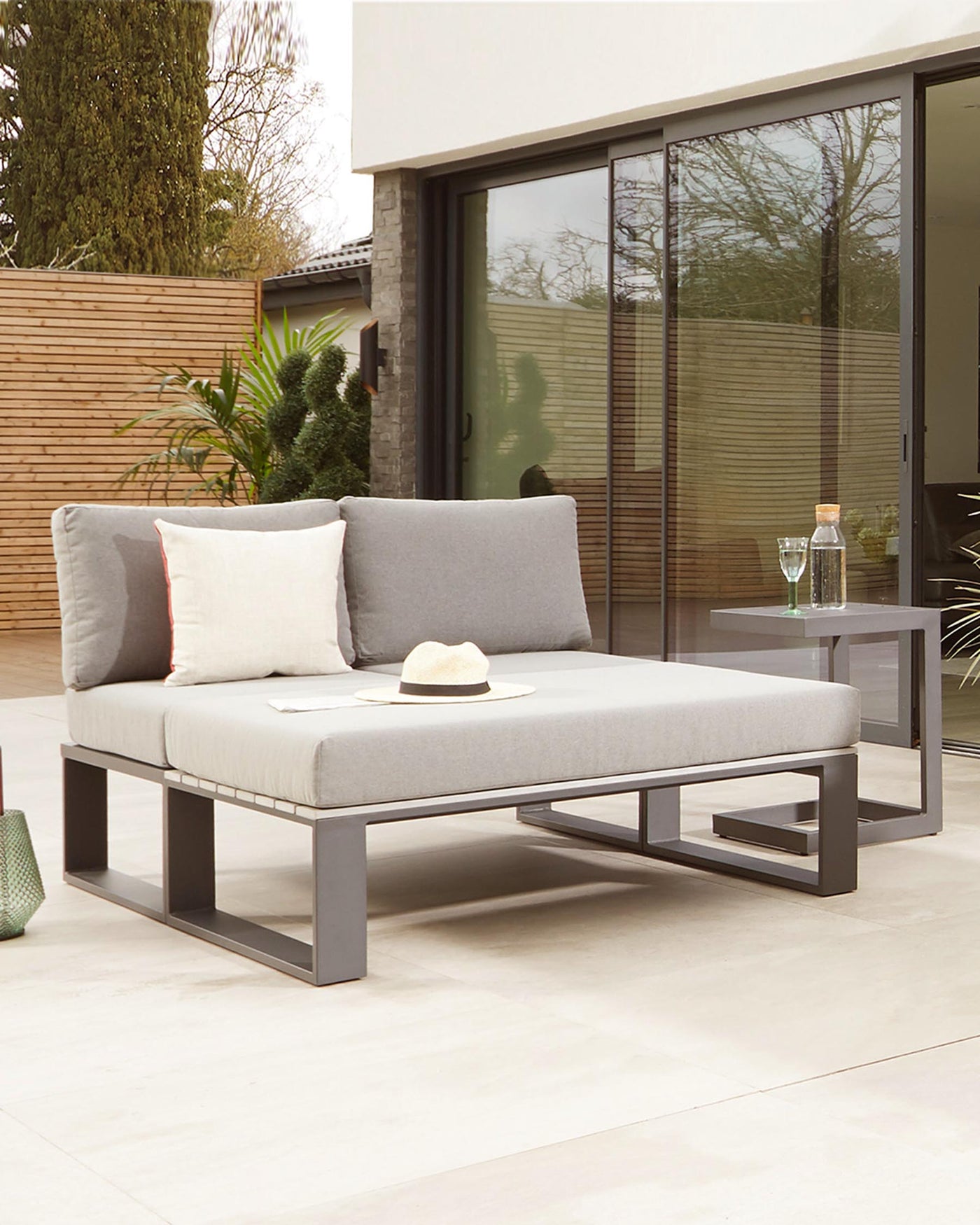Modern outdoor lounge set featuring a sleek, grey metal frame chaise lounge with light grey cushions and complementary throw pillows, paired with a matching grey metal frame side table with a clear glass top.