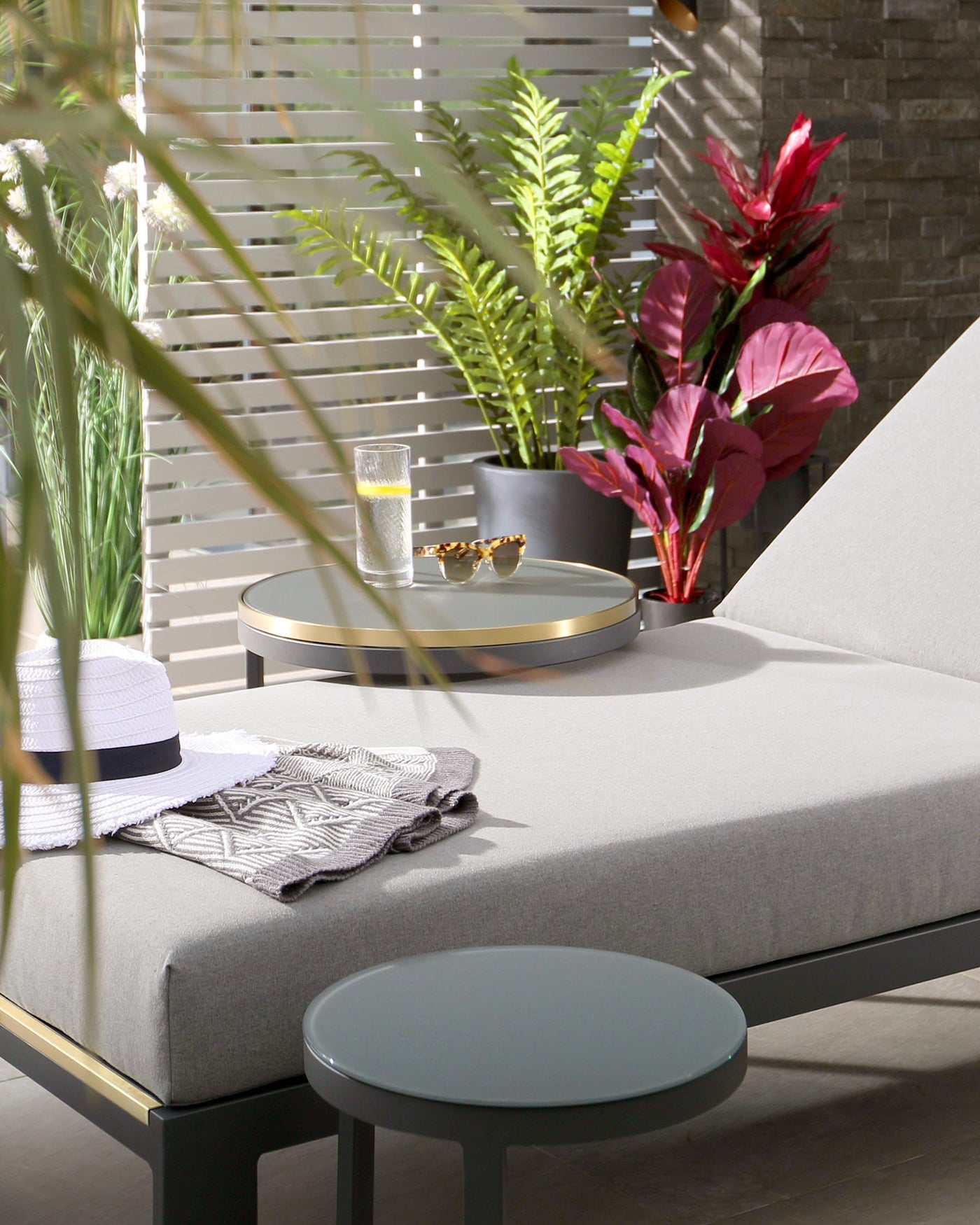 Modern outdoor furniture featuring a sleek, grey chaise lounge with black metal framing and a side table with a round, pale green top and black base. Additional circular centrepiece table with a golden rim and a two-tiered design, with the top displaying a glass of water and sunglasses. The setting is complemented by decorative plants, providing a fresh, natural ambiance.